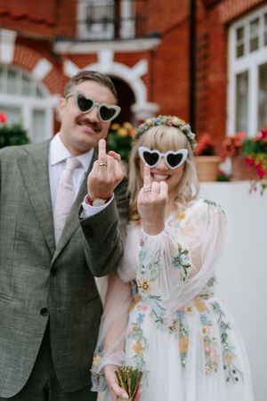 A couple wearing heart-shaped sunglasses make rude gestures at the camera in front of a brick building.