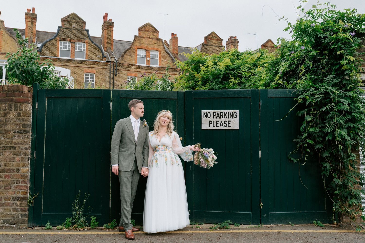 A couple in formal wedding attire walks together near a brick wall and green fence in an outdoor garden setting.