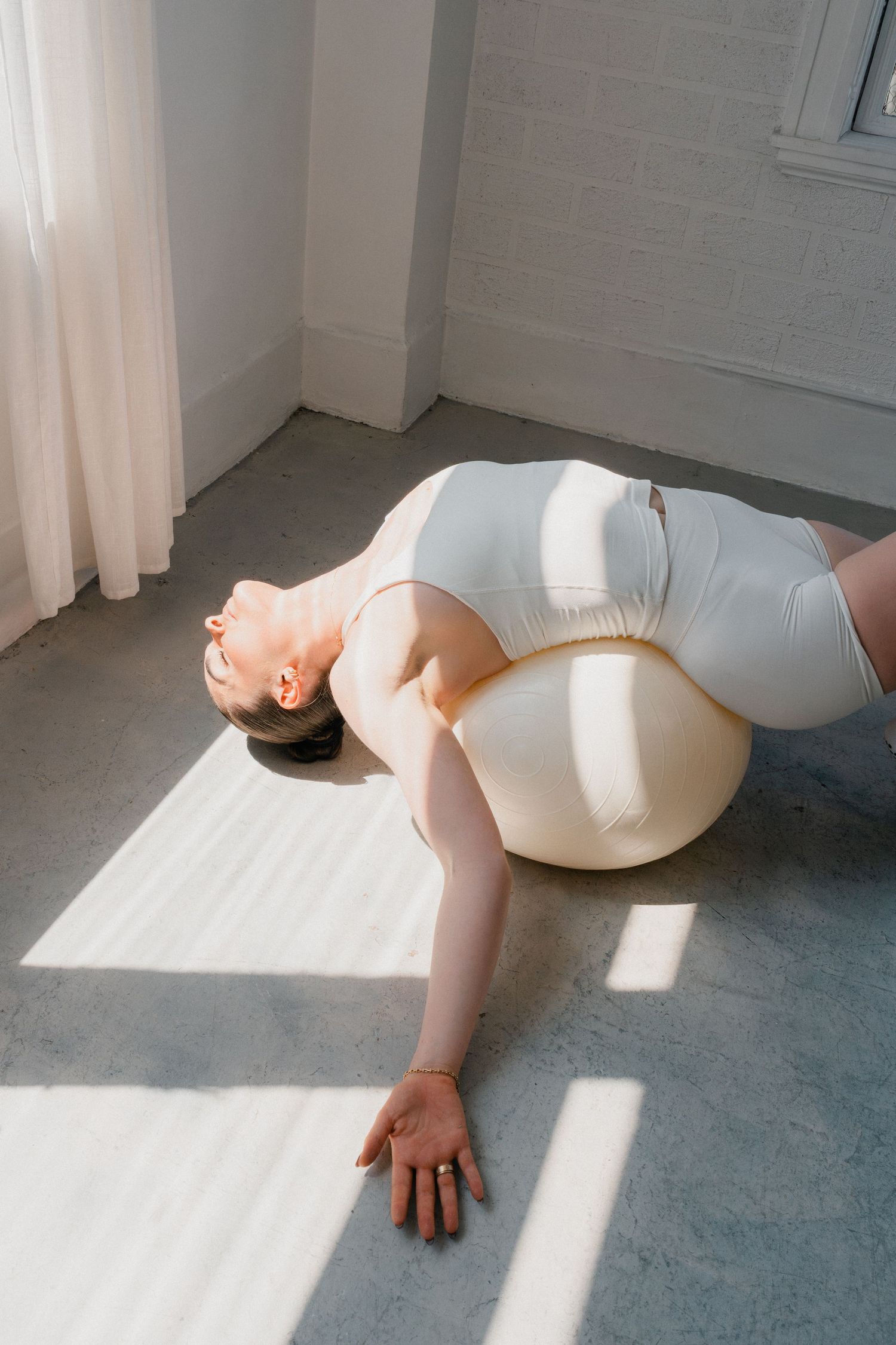 Person in white activewear laying on yoga ball in sunlit room with white brick walls during exercise or stretching session.