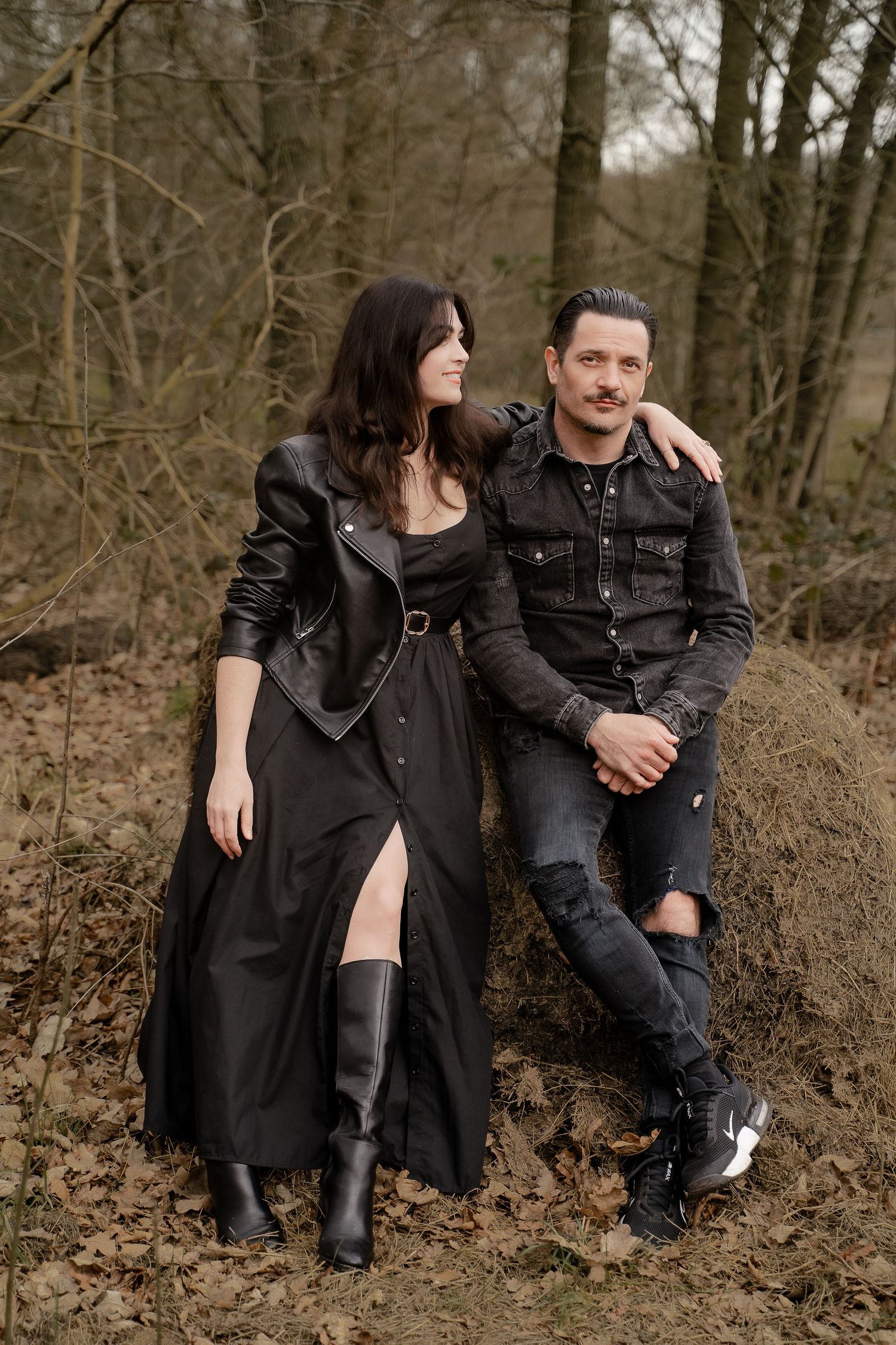 A couple sits together on a mossy log in a quiet woodland area near Measham, dressed in relaxed dark tones, sharing a moment of calm during