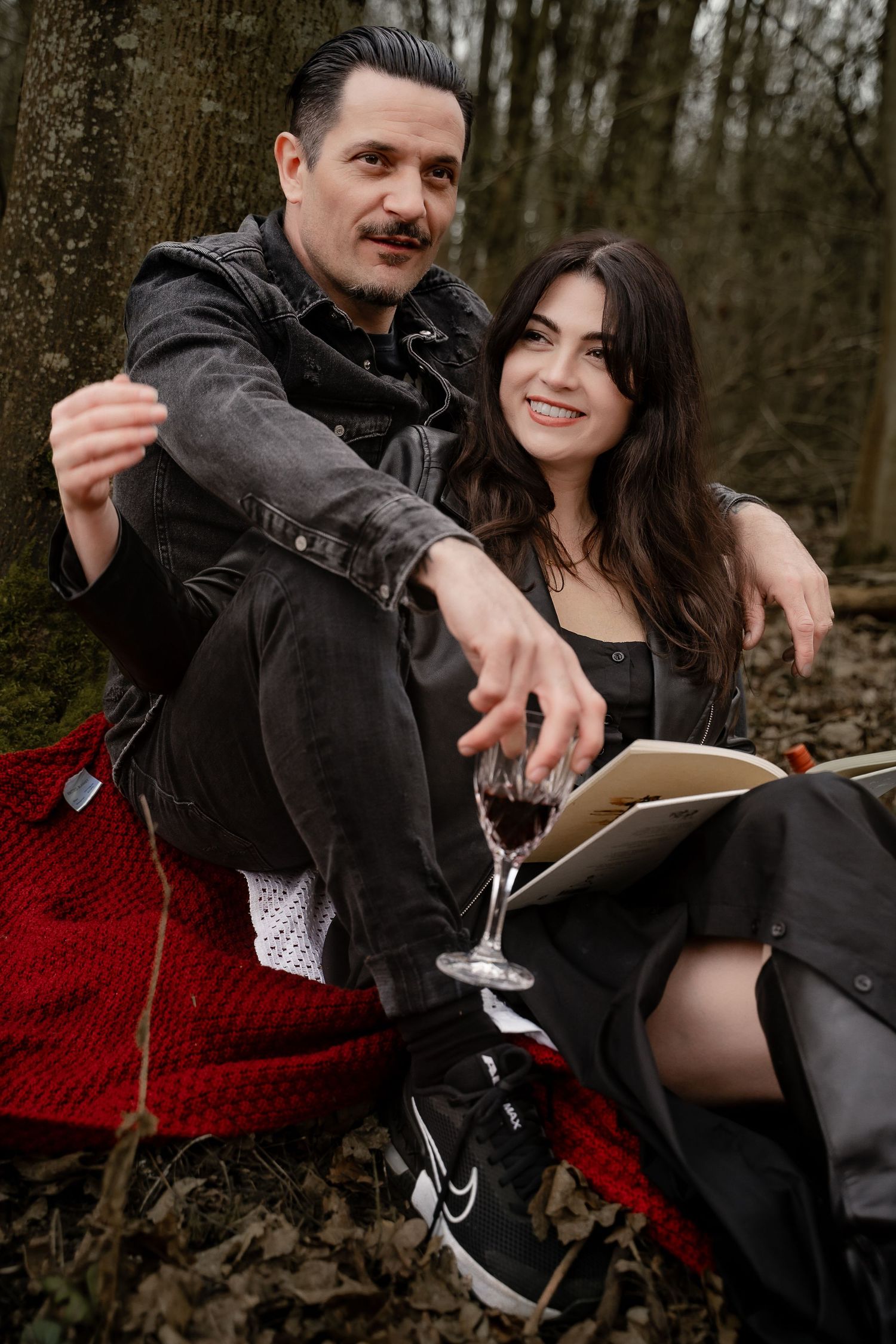 A couple sits close together at the base of a tree in Measham woodland, smiling softly with arms wrapped around each other — a cosy, candid