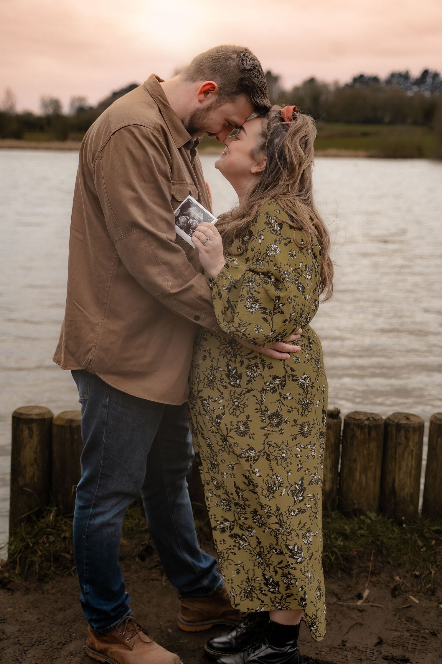 A couple stands forehead to forehead by the water, smiling and holding their baby scan photo during a maternity session near Measham. A warm