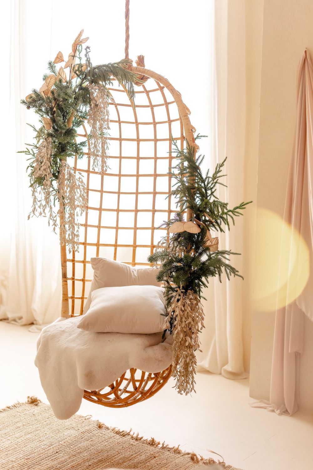 Cozy hanging rattan chair decorated with greenery and draped with soft cushions and throw blanket in a sunlit room.