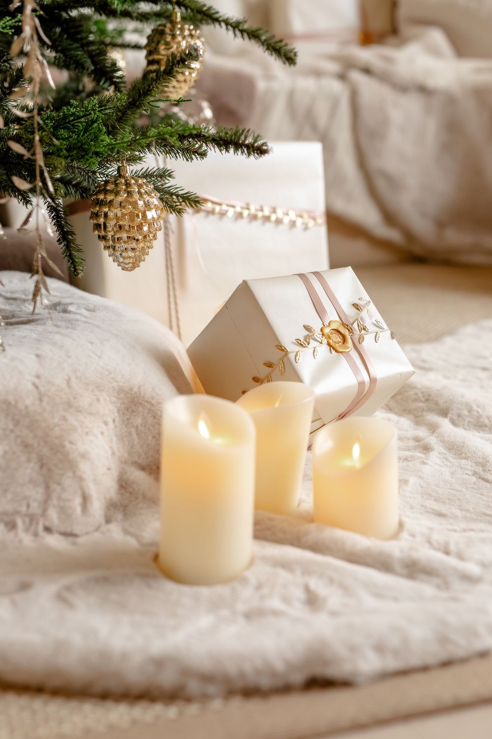 Christmas decor featuring lit candles, pine branches with gold ornaments, and wrapped presents on a white textured blanket.