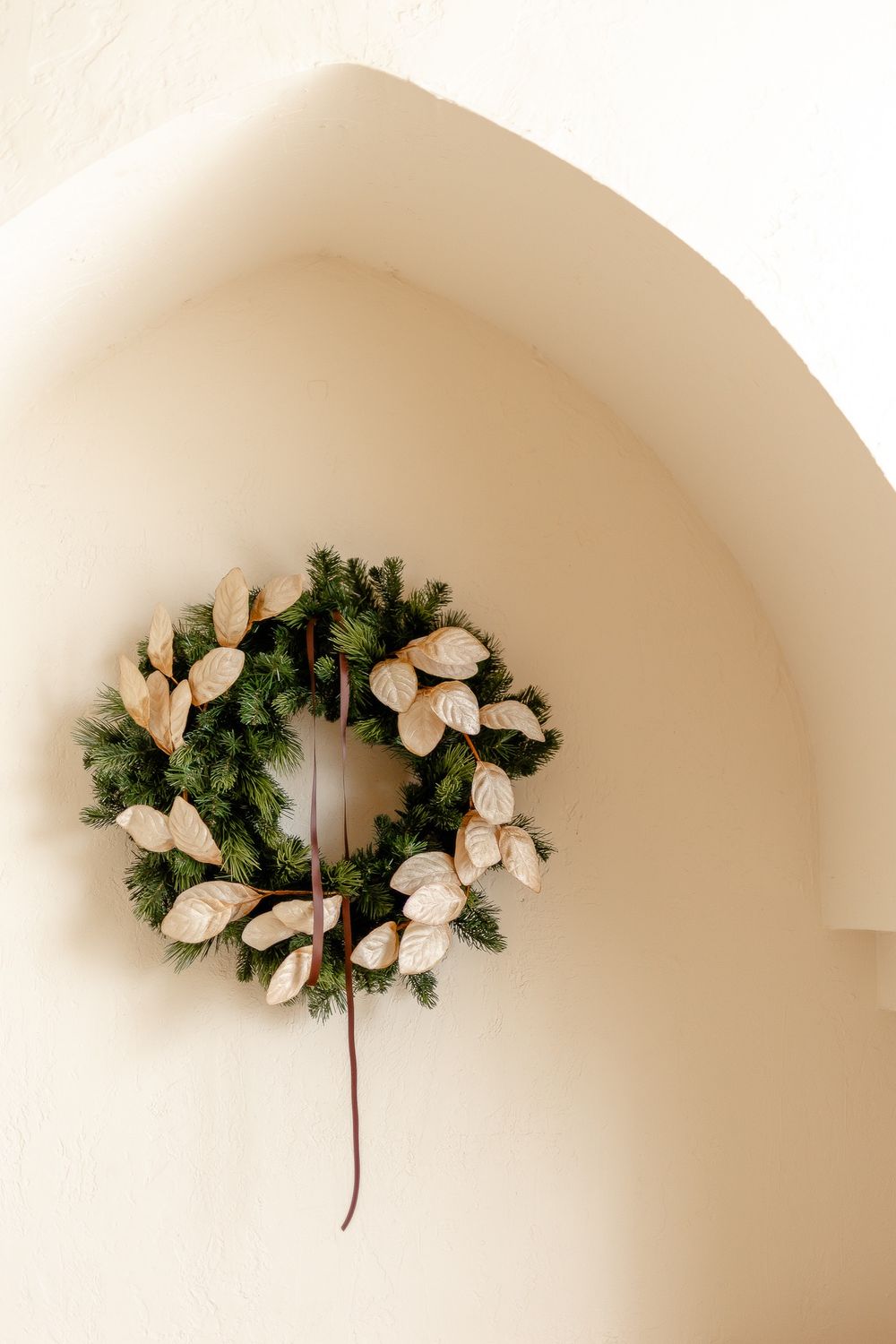A minimalist Christmas wreath with white poinsettias and evergreen branches hangs on a cream-colored wall.