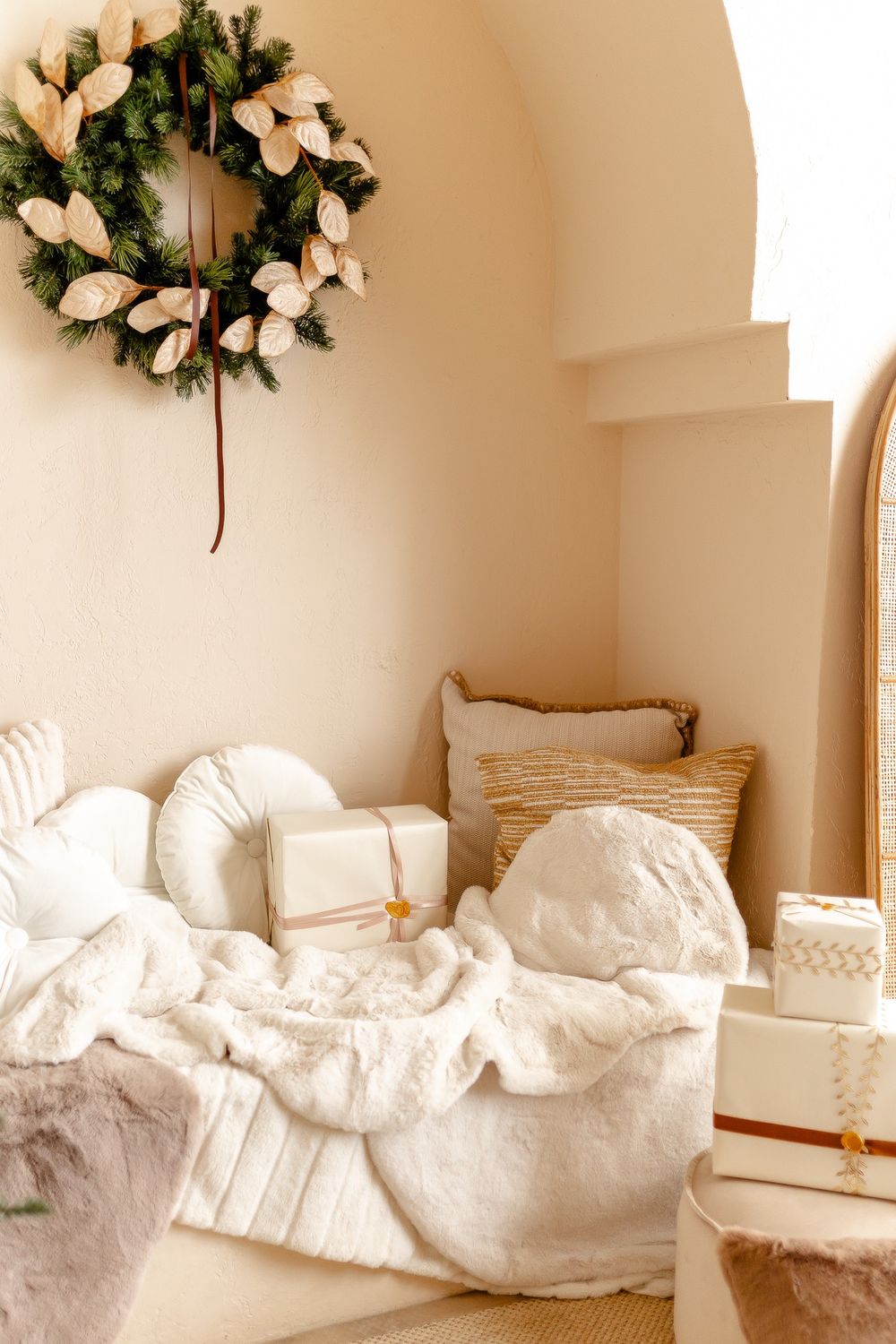 Cozy white bedroom corner with plush bedding, decorative wreath, and neutral decor in warm lighting.
