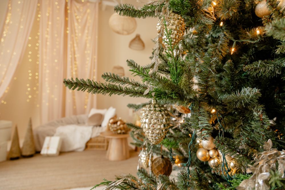 A Christmas tree adorned with gold ornaments and lights stands elegantly in a warm-lit corner with white curtains.