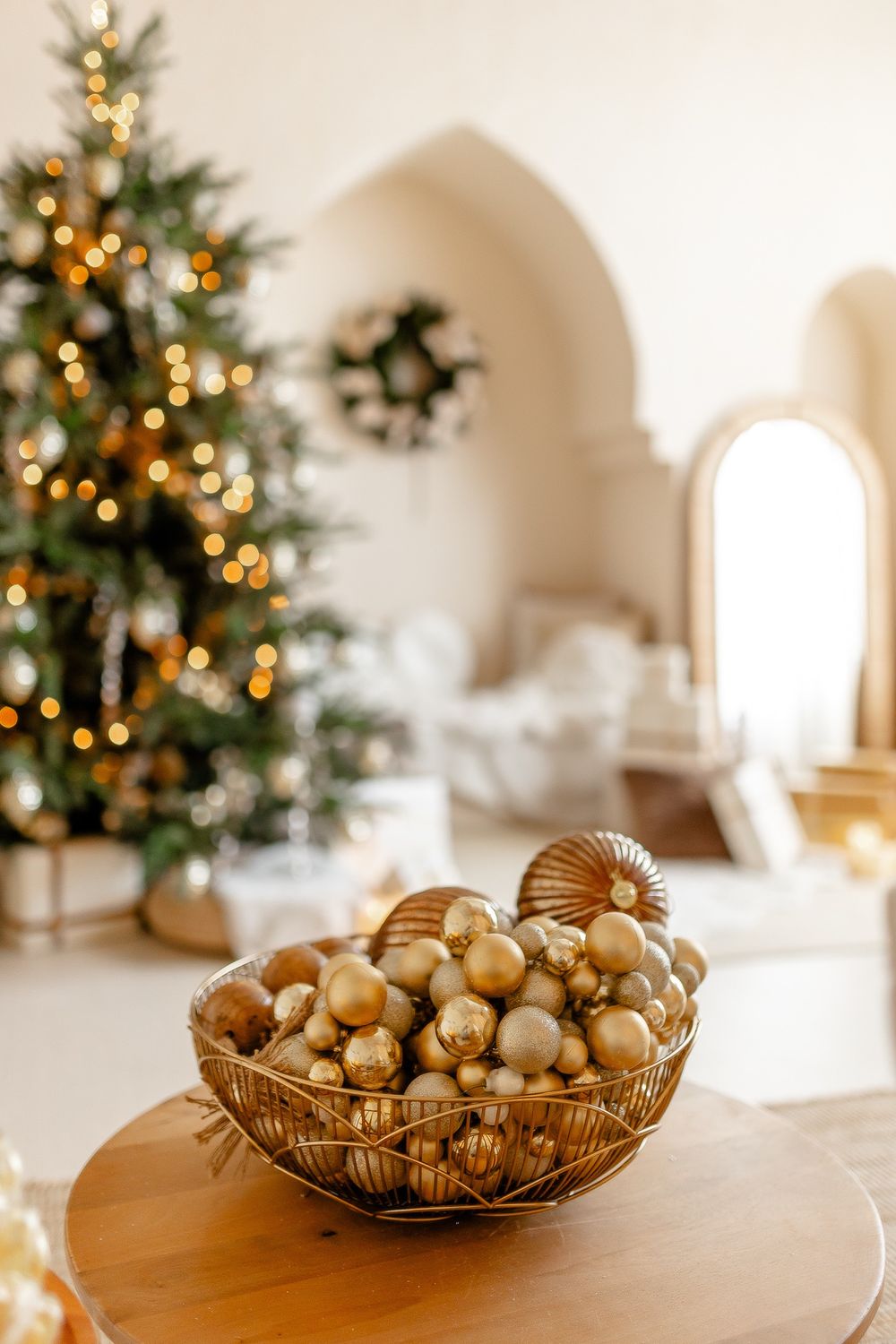 Glass bowl filled with golden Christmas ornaments on wooden table with decorated tree in soft focus background.
