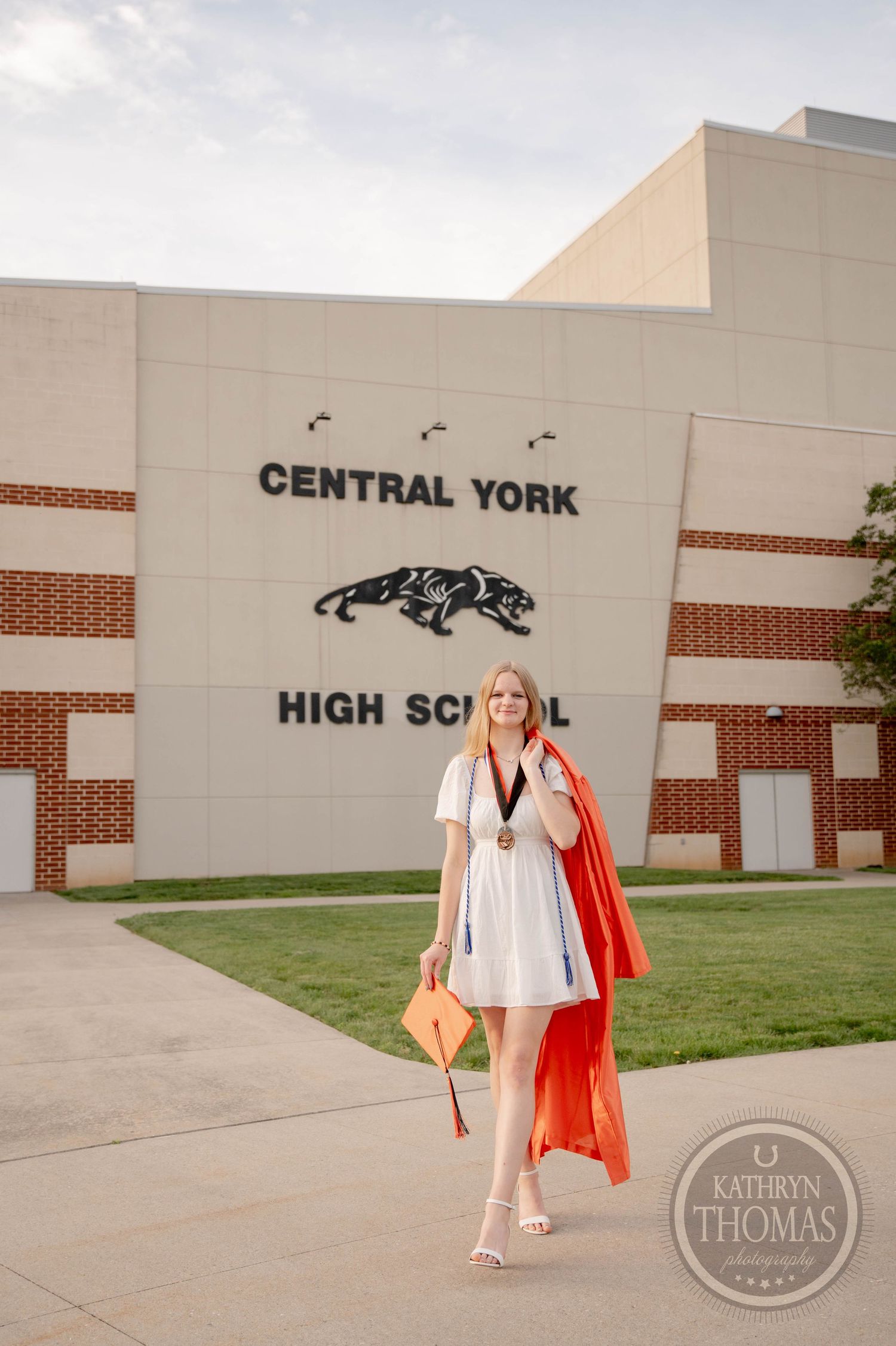 Graduate in white dress with orange sash walks past Central York High School sign during sunny outdoor photo session.