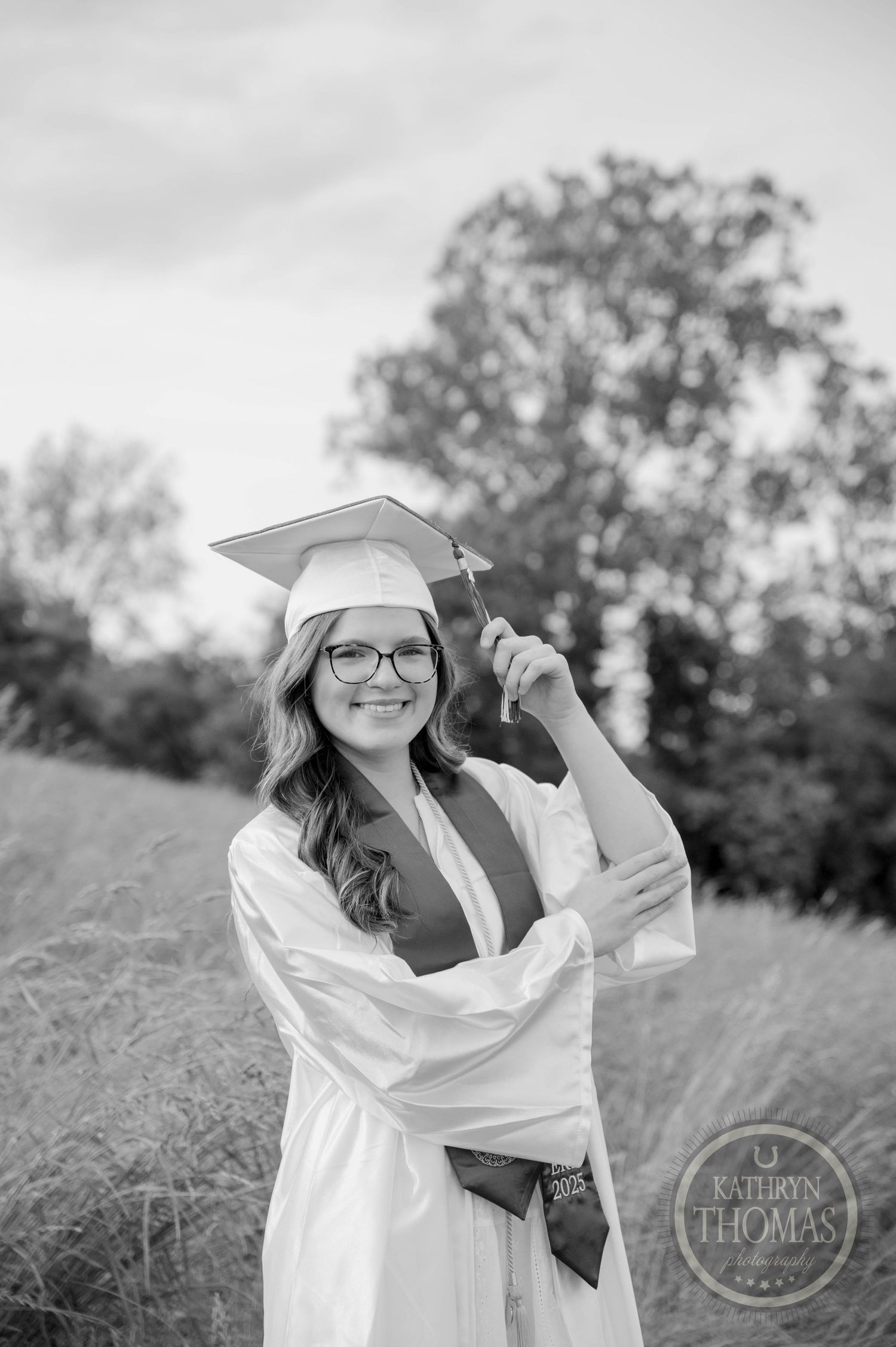 Black and white graduation photo of student in cap, gown and glasses posing outdoors in a grassy field.