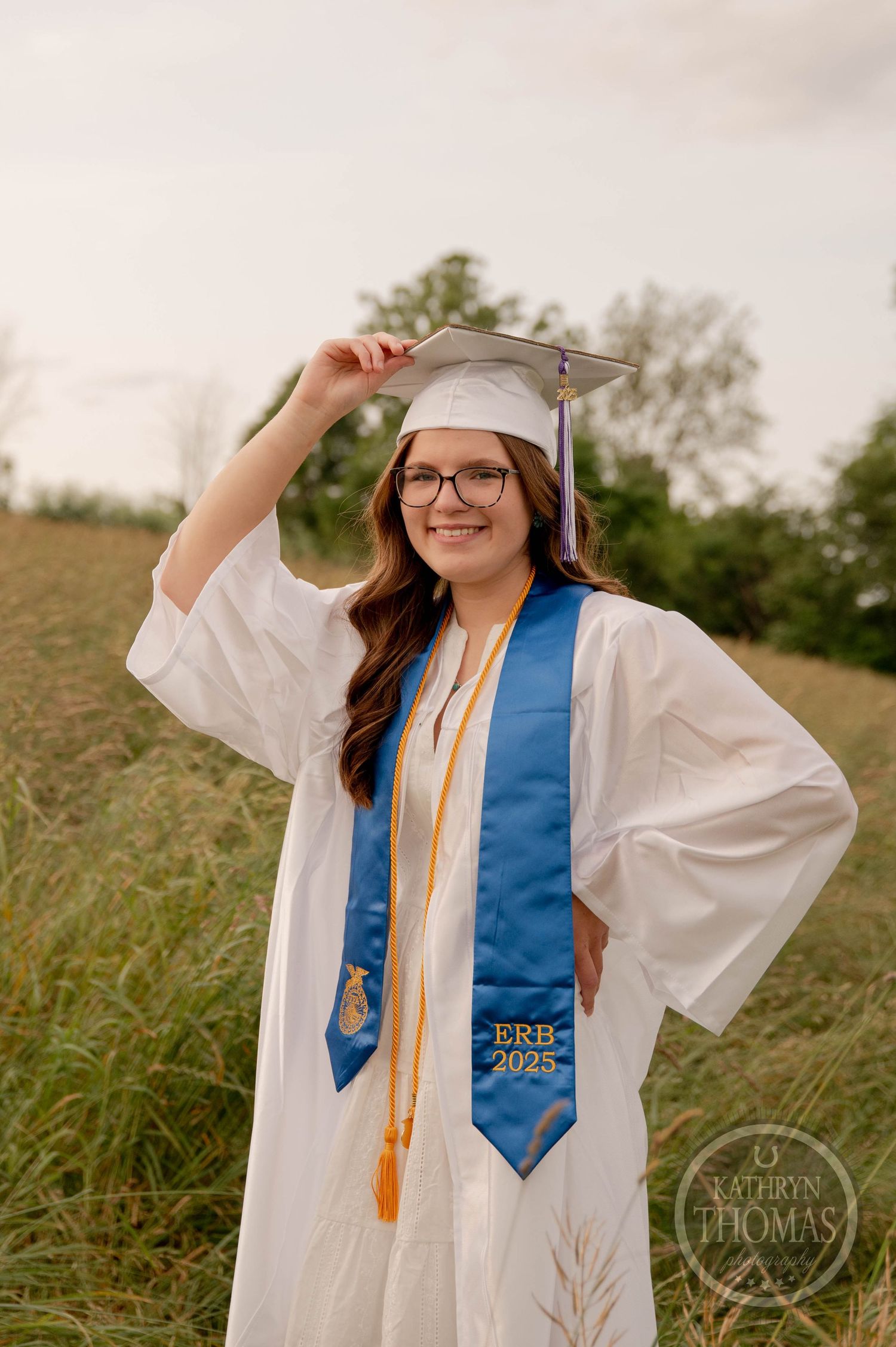 Graduate in white cap and gown with blue sash celebrates achievement in outdoor field setting with joyful expressions.