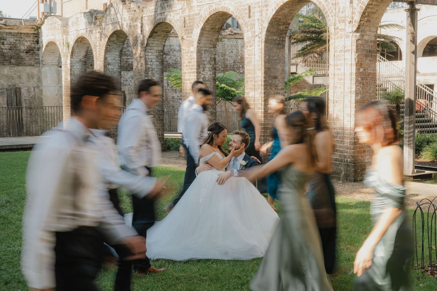 Motion blur captures wedding party dancing and celebrating under stone archways in a garden setting.