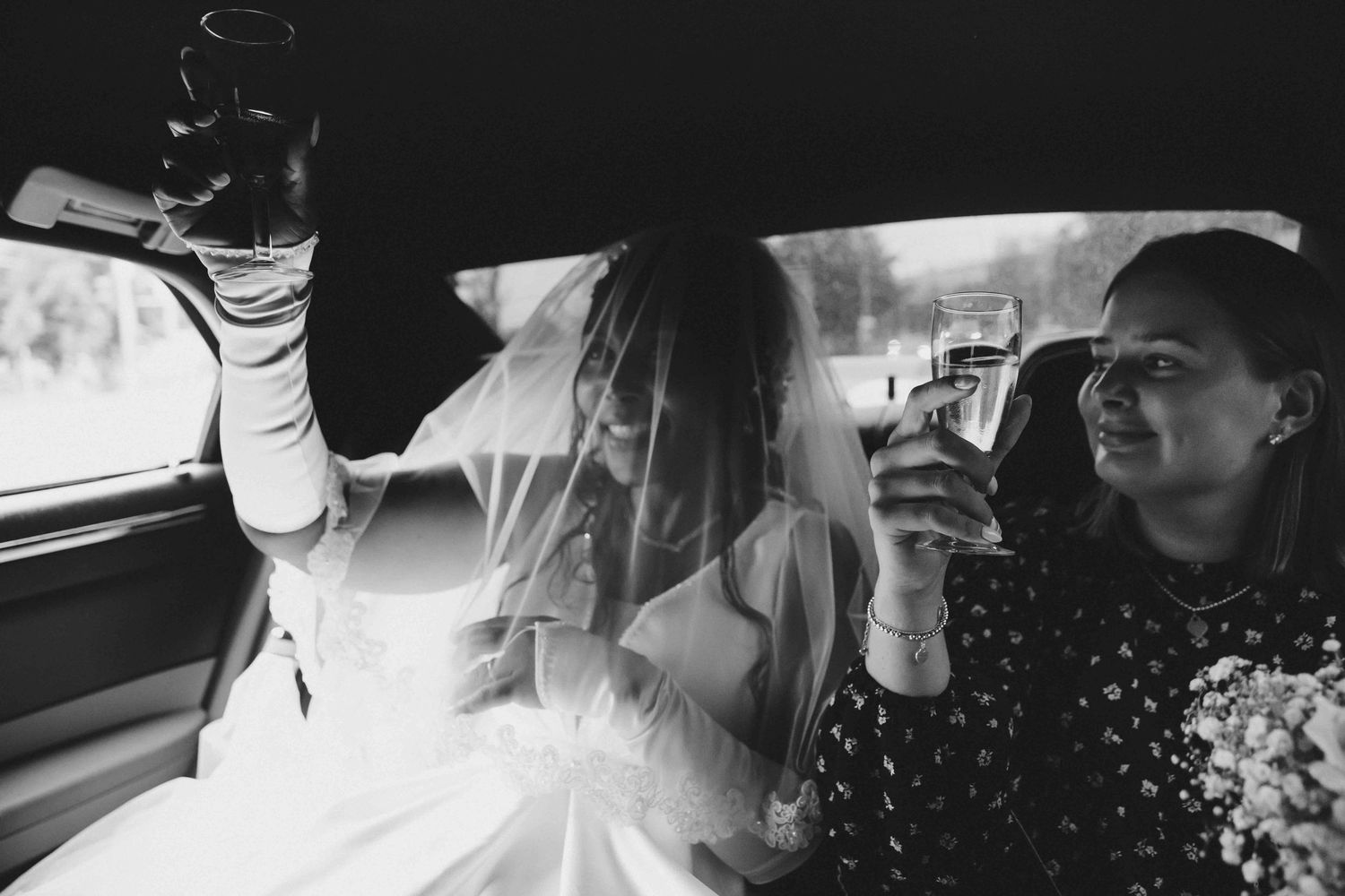 Two people clink glasses while riding in a car, one in a white veil, captured in black and white photography.