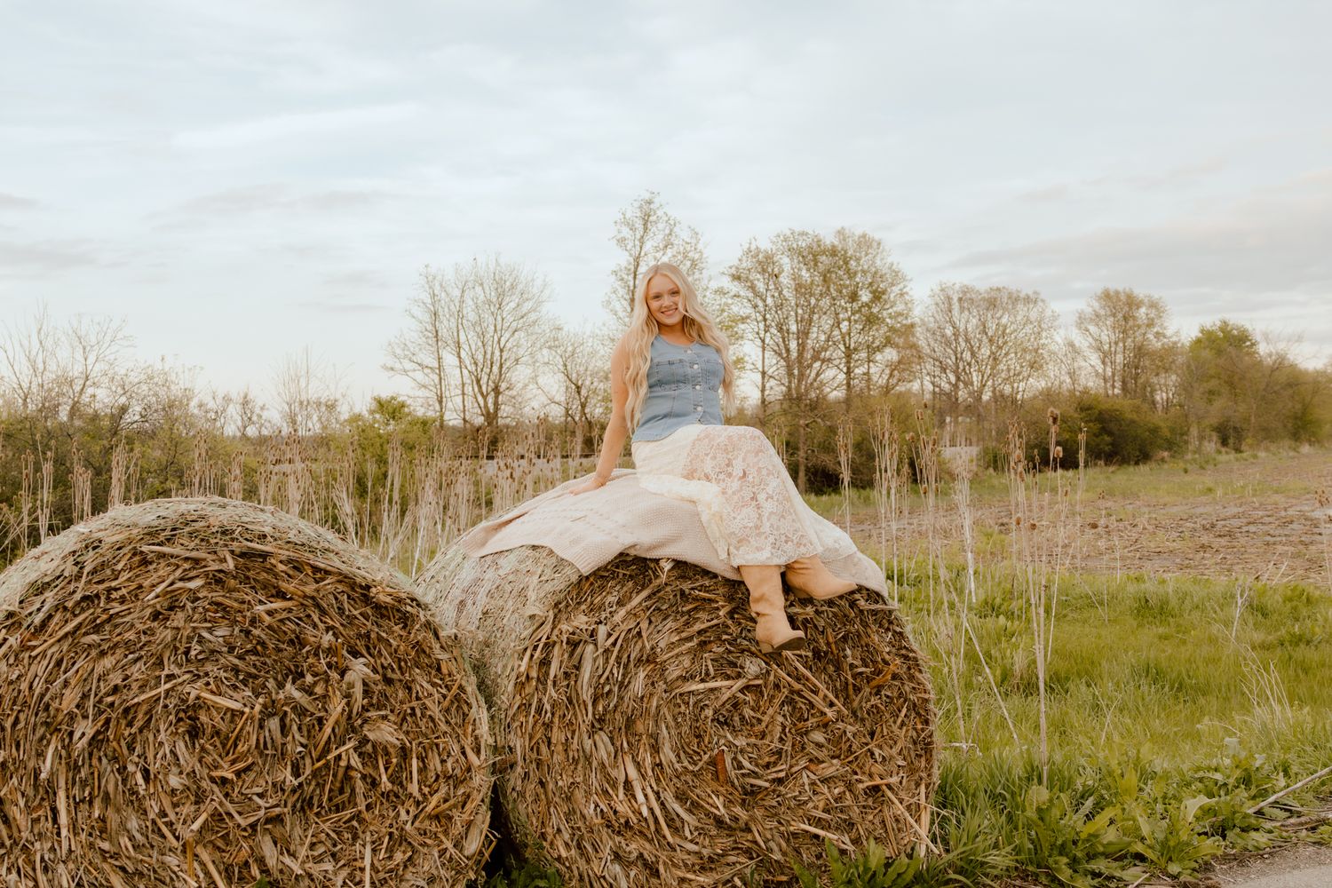 Young blonde girl sitting atop large round hay bales in an open farm field.