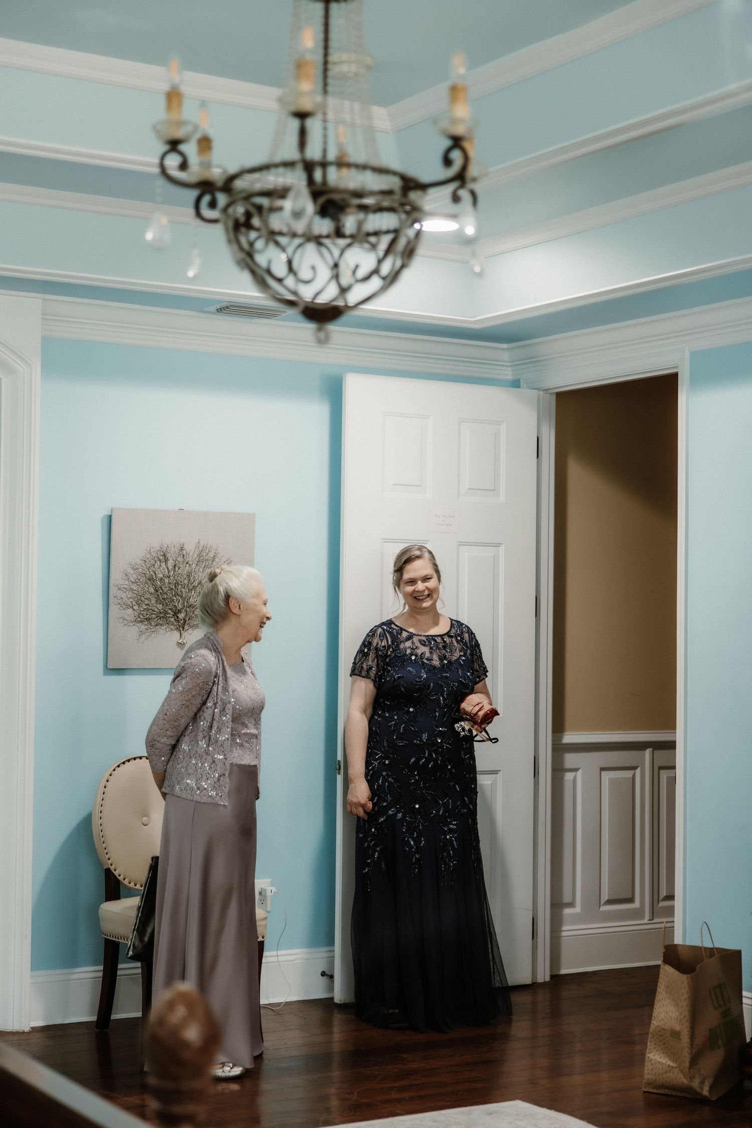 Two people in formal evening attire share a joyful moment in an elegant blue-walled room with a vintage chandelier.
