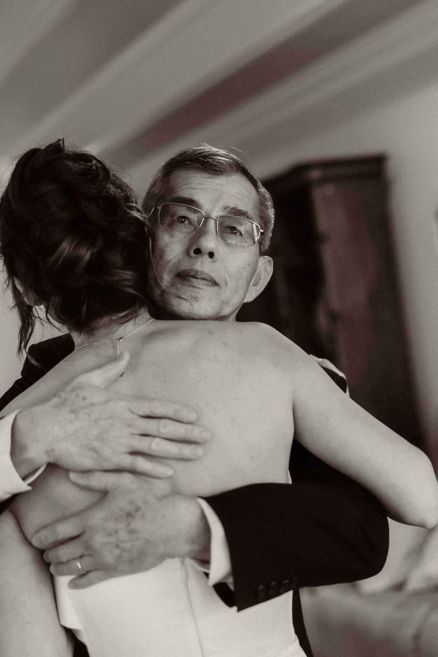 An intimate black and white photograph captures an emotional embrace between two people at what appears to be a wedding.