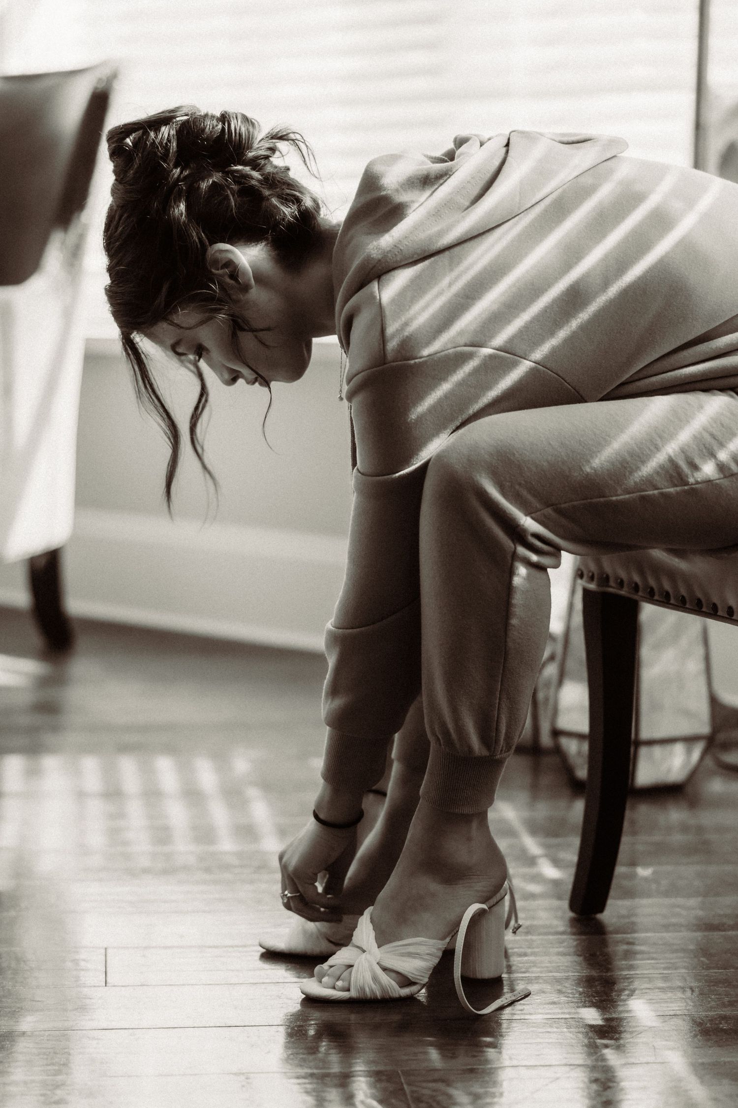 Person in casual attire leaning forward on a chair while adjusting their footwear on a hardwood floor.
