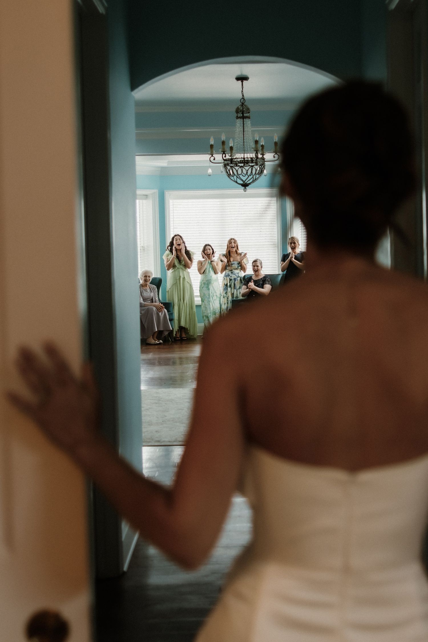 Bridesmaids in mint green dresses react with joy as they see someone in a white dress through an arched doorway.