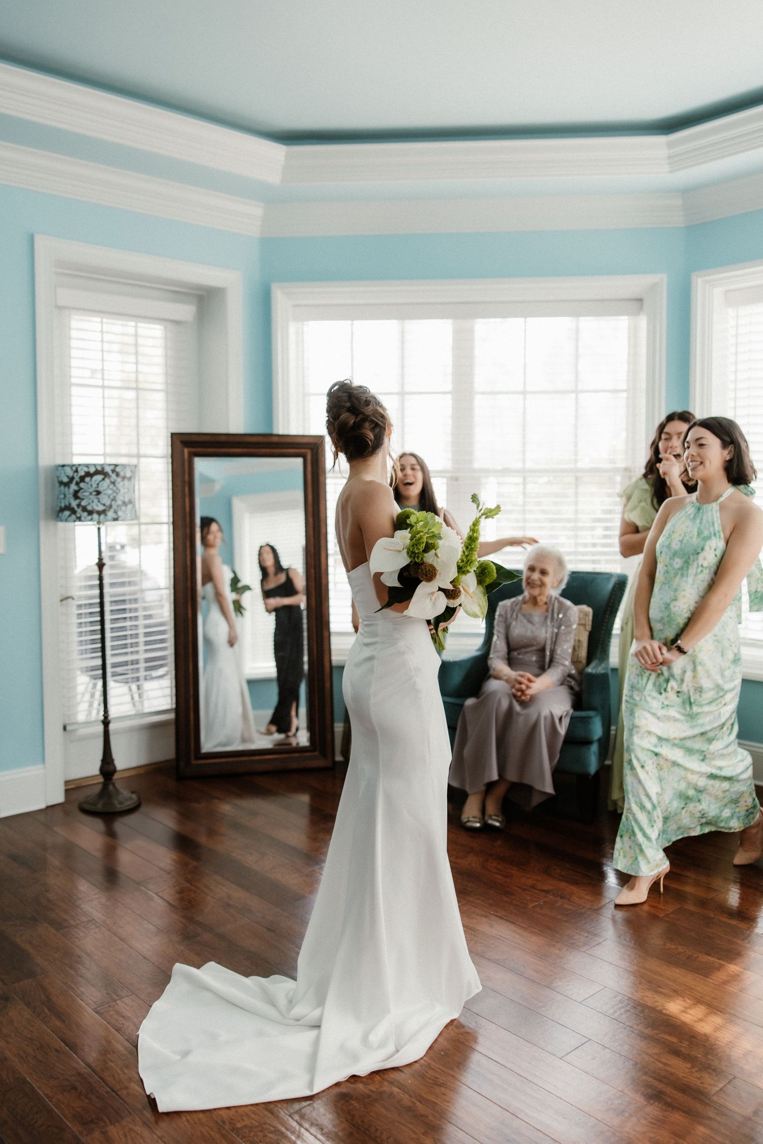 A bride in a sleek white gown admires her reflection in a full-length mirror while others watch in a light blue room.