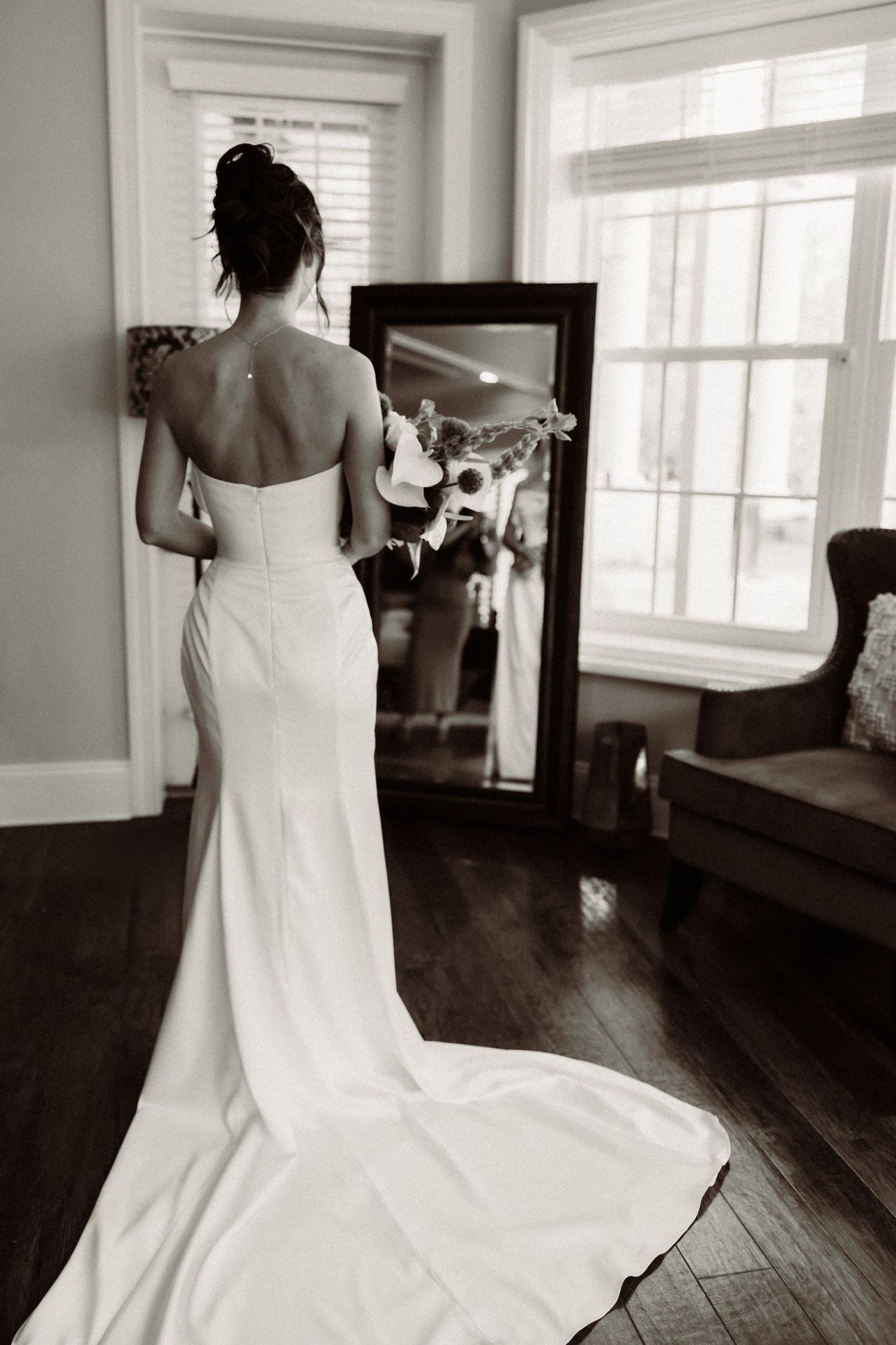 A black and white photo of a bride in an elegant fitted wedding gown with a train looking at her reflection.