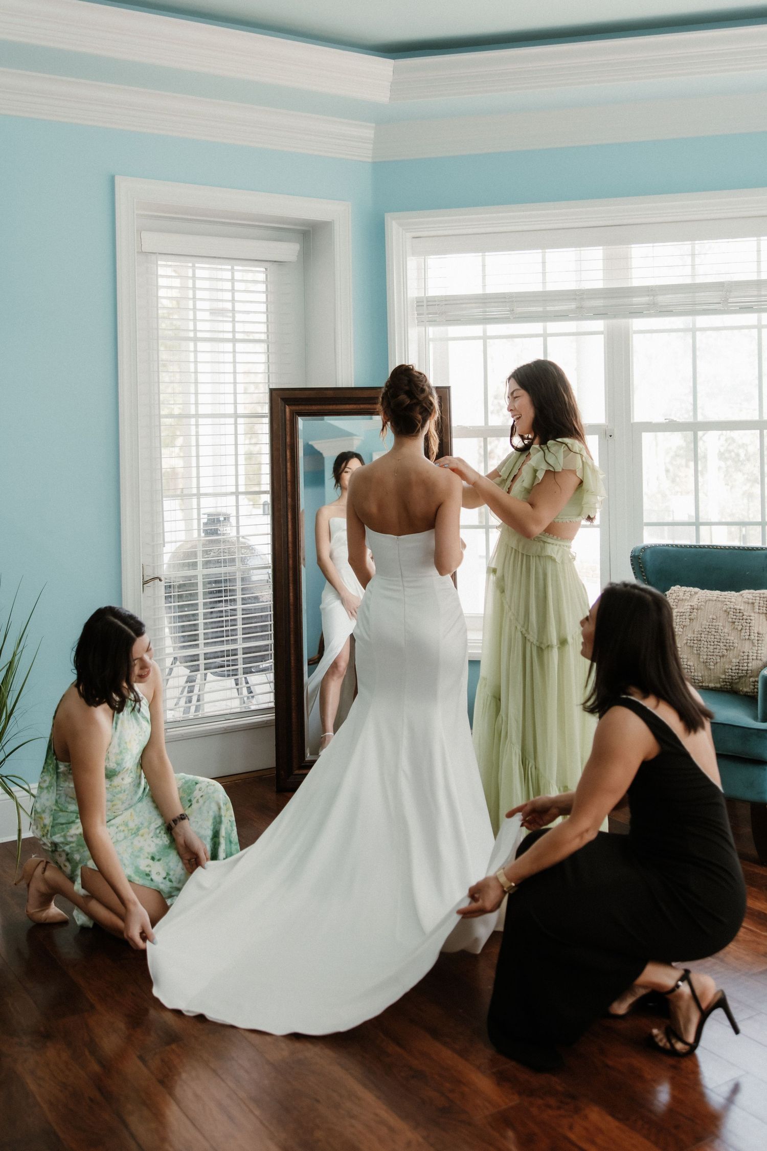 Wedding preparations in a blue room as attendants help adjust a bride's flowing white gown.
