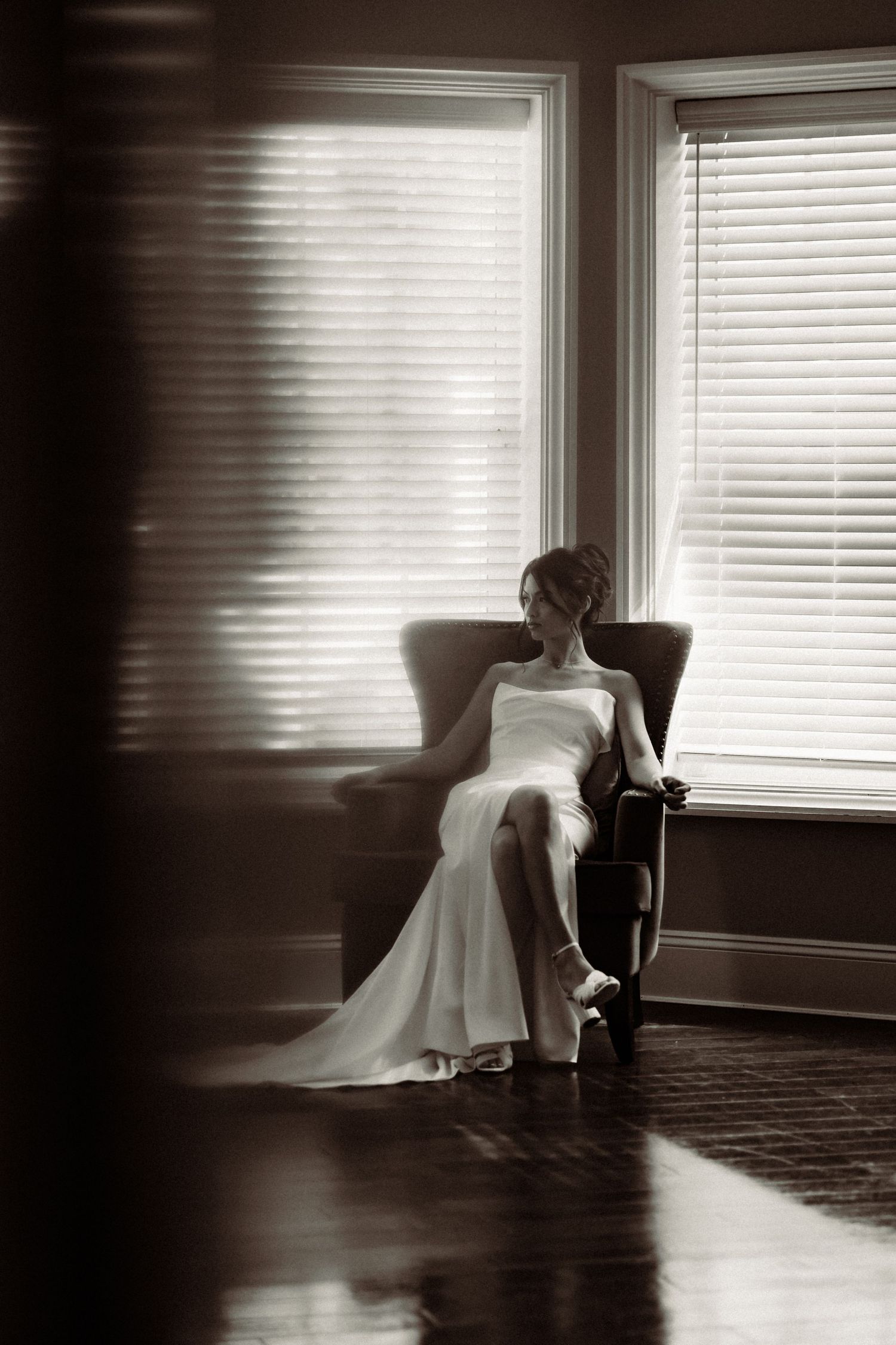 A bride in a white gown sits elegantly in a chair by windows with sunlight streaming through horizontal blinds.