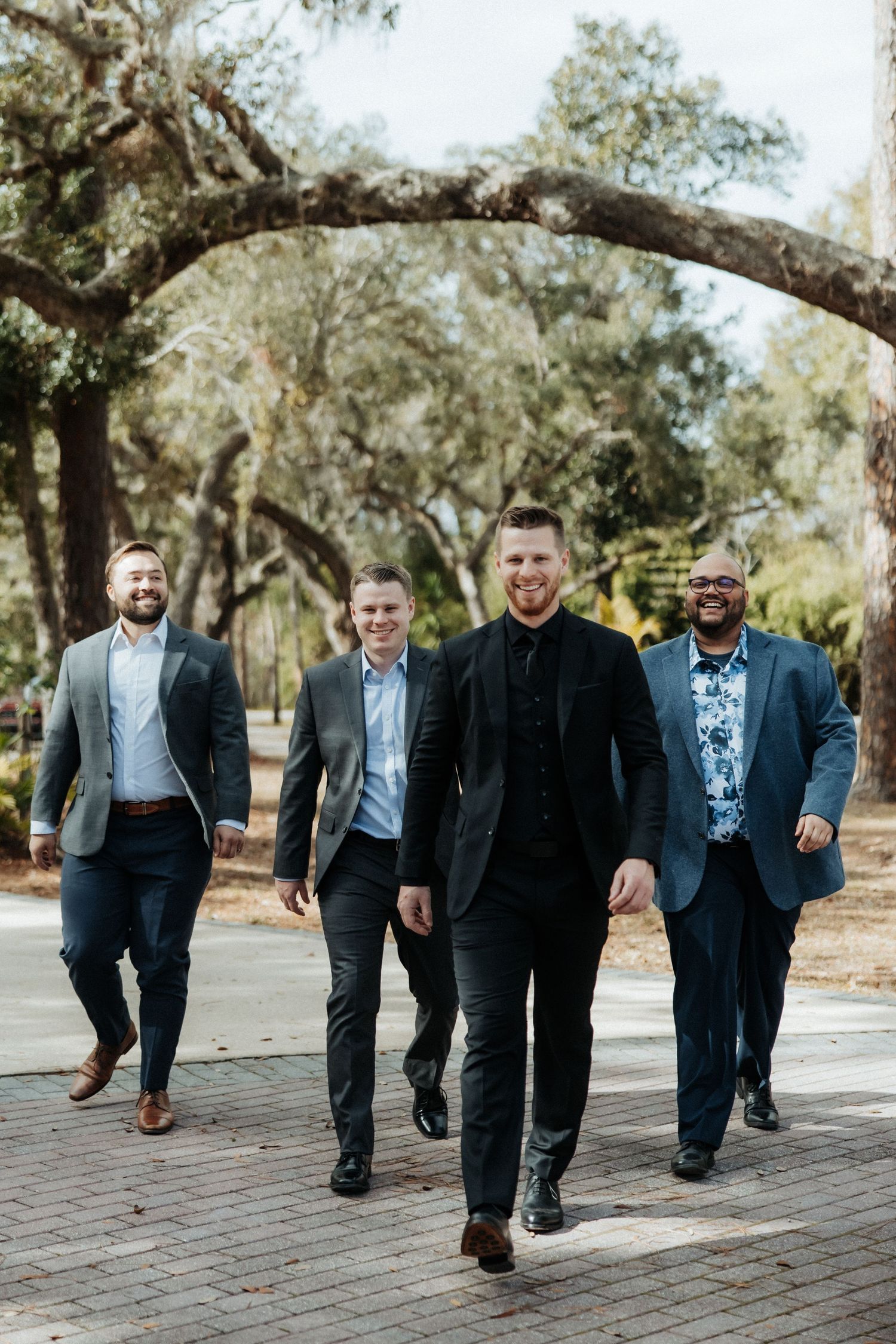A group of groomsmen walking confidently on a tree-lined path in matching suits and blazers.