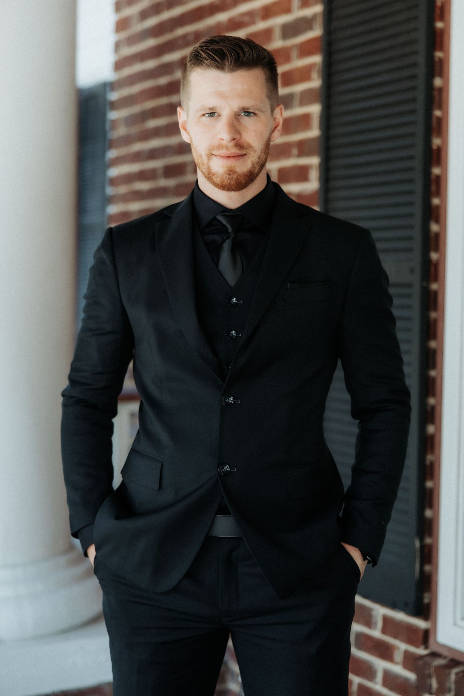 A fashionable three-piece black suit with tie is modeled against a brick and white column backdrop.