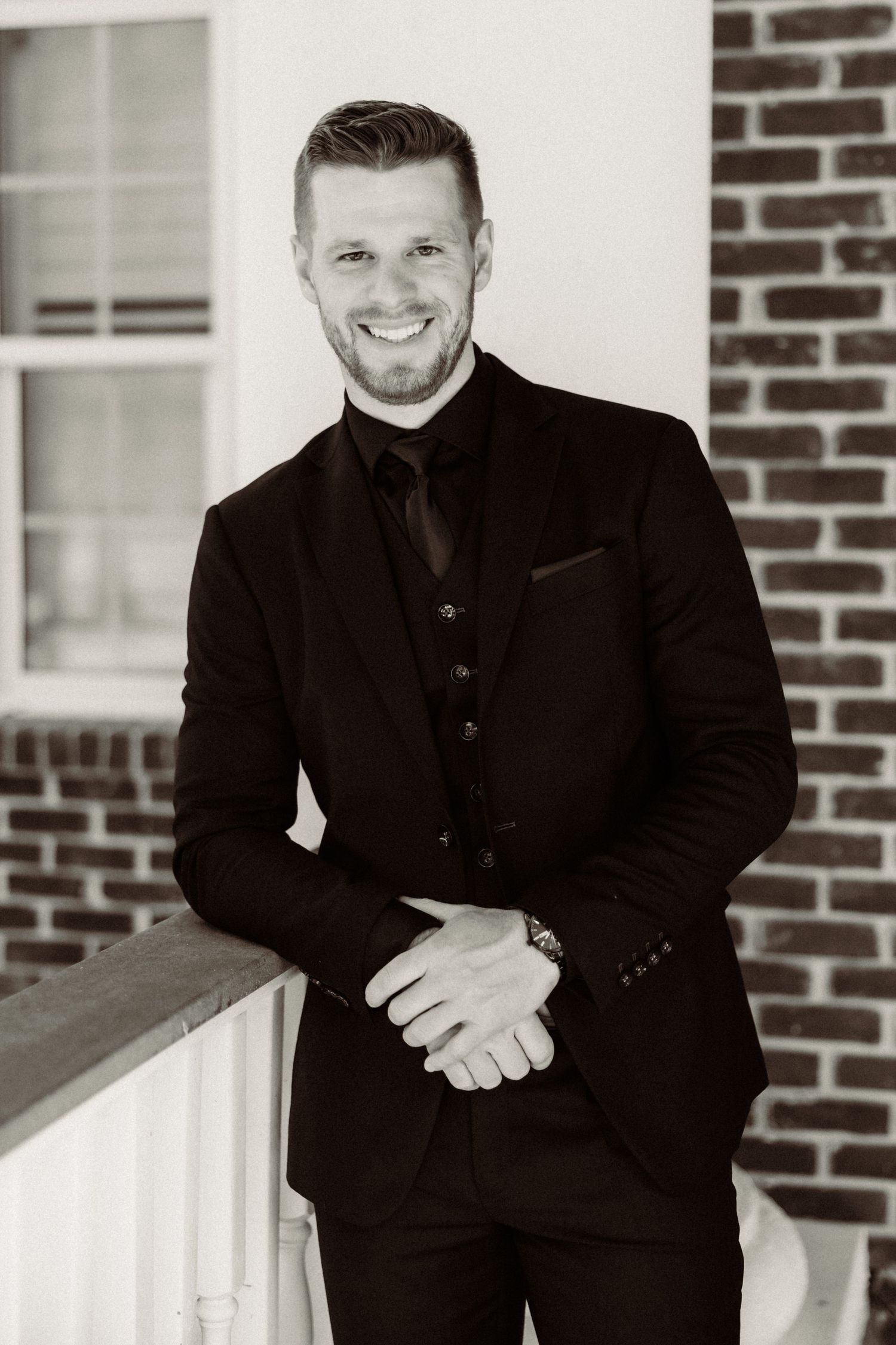 A smiling person wearing a black formal suit and tie poses against a brick wall in a black and white portrait.