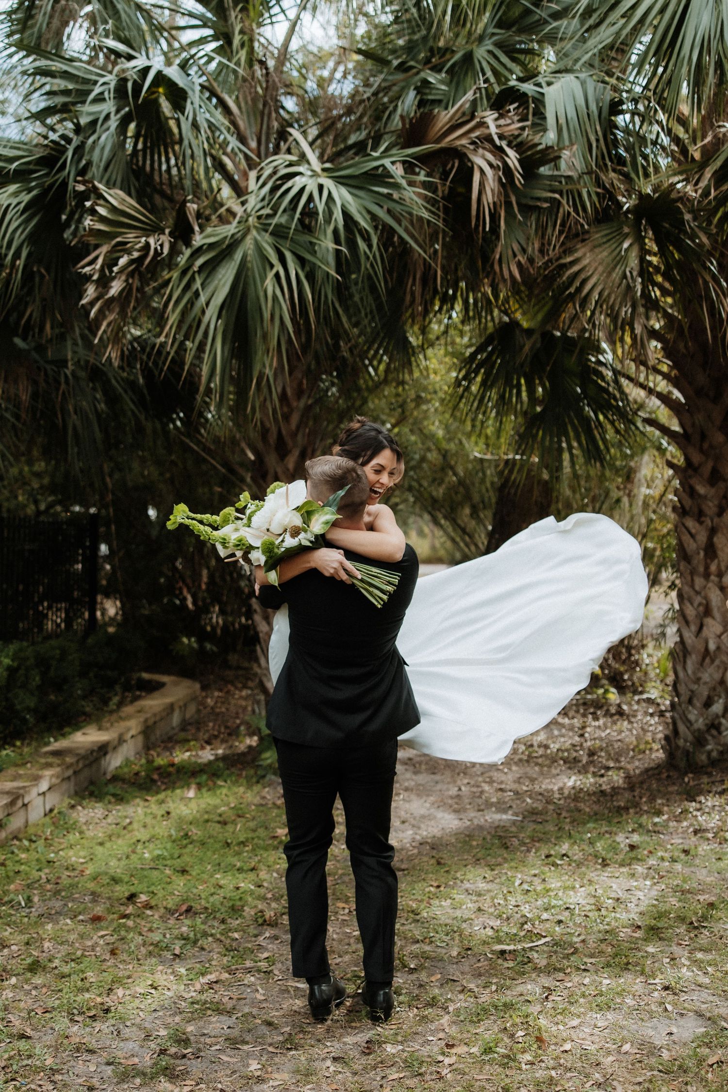 A romantic wedding photo under palm trees shows an embrace with flowing white dress fabric billowing in the breeze.