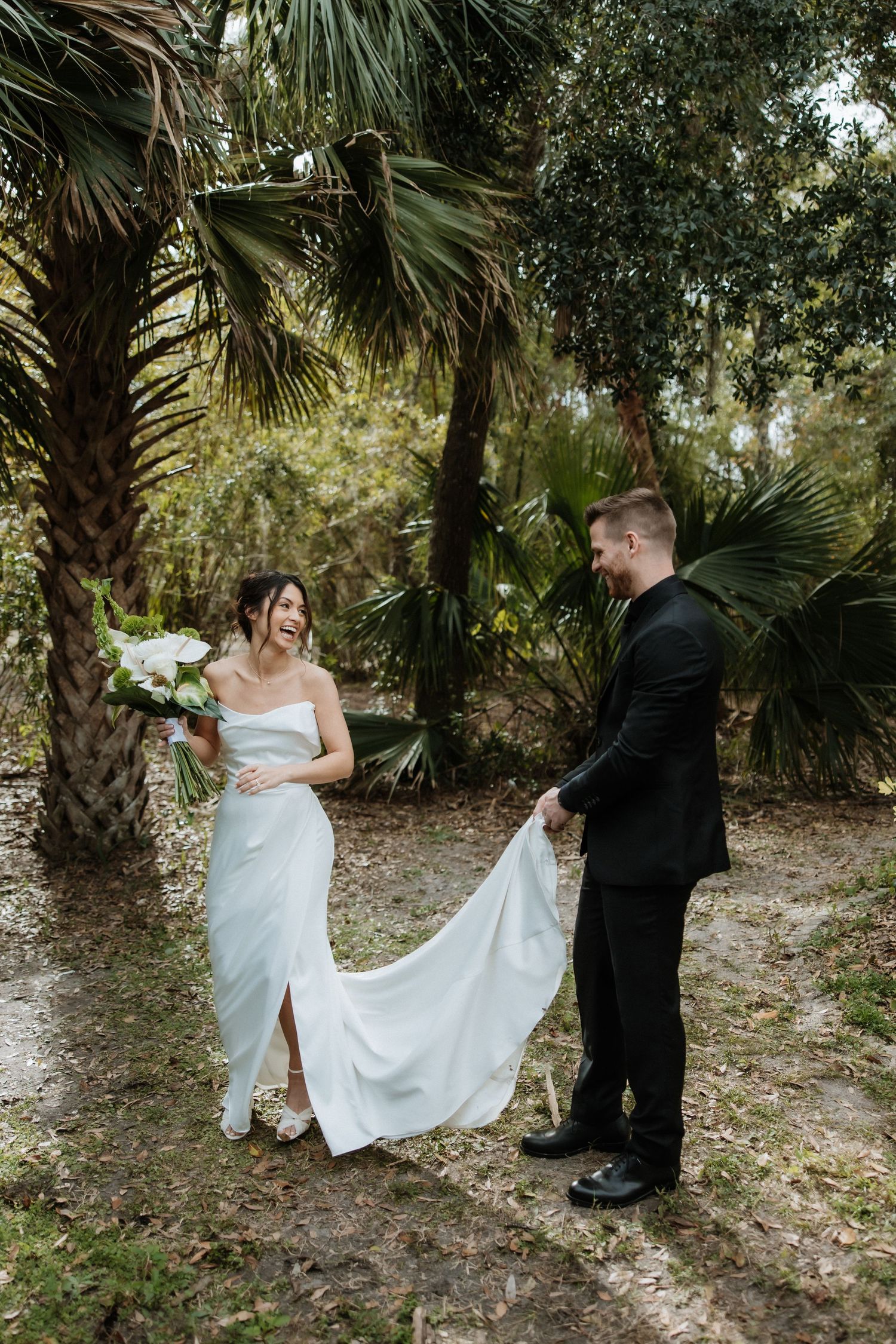 A wedding couple walks among palm trees in an intimate outdoor setting with sunlight filtering through.