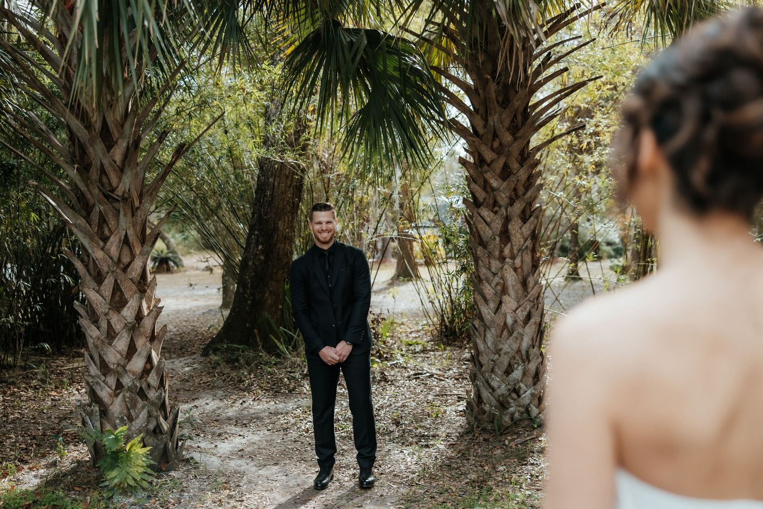 A romantic first look captured between palm trees on a sandy beach pathway.