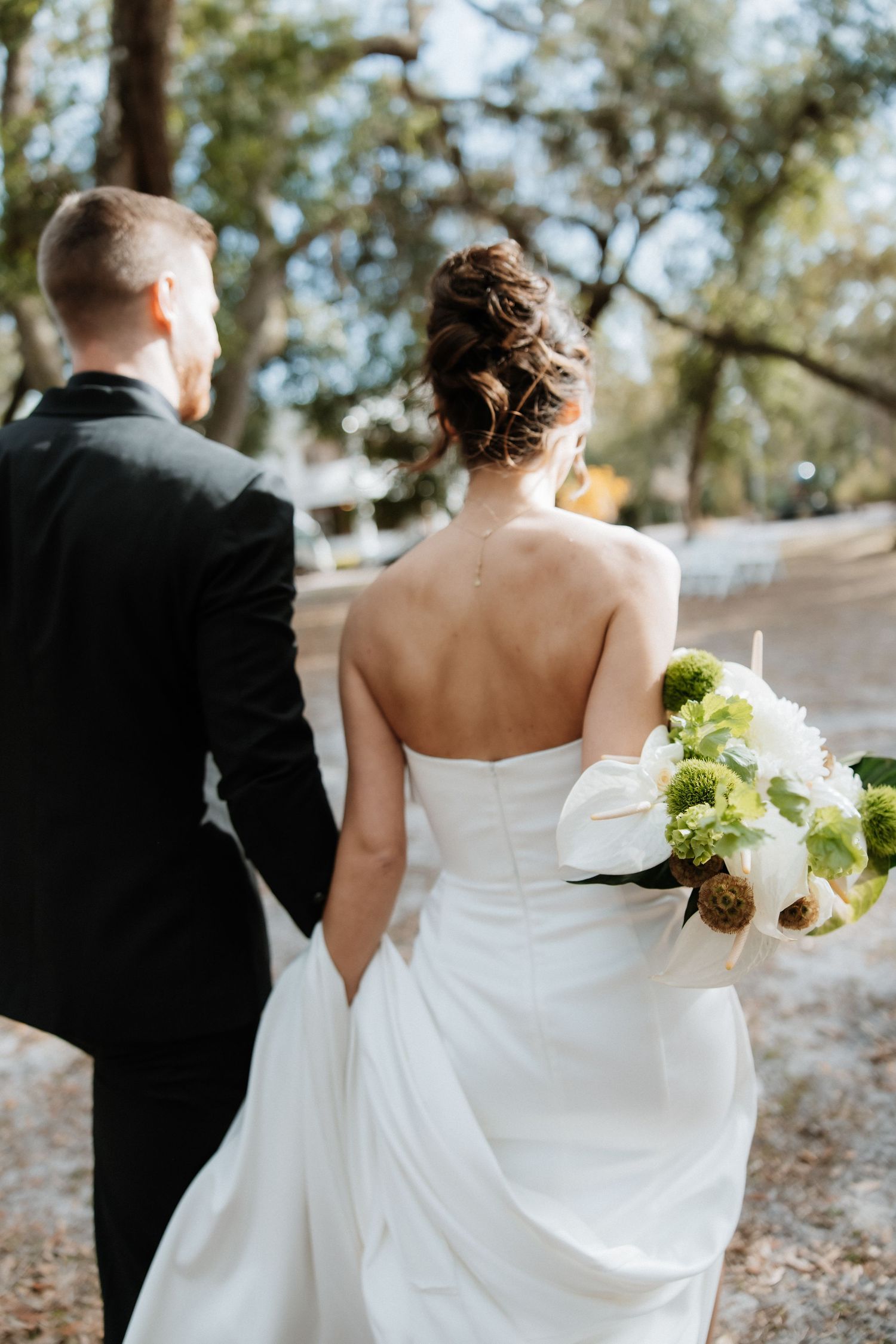Couple in wedding attire walking through a scenic outdoor path with white florals.