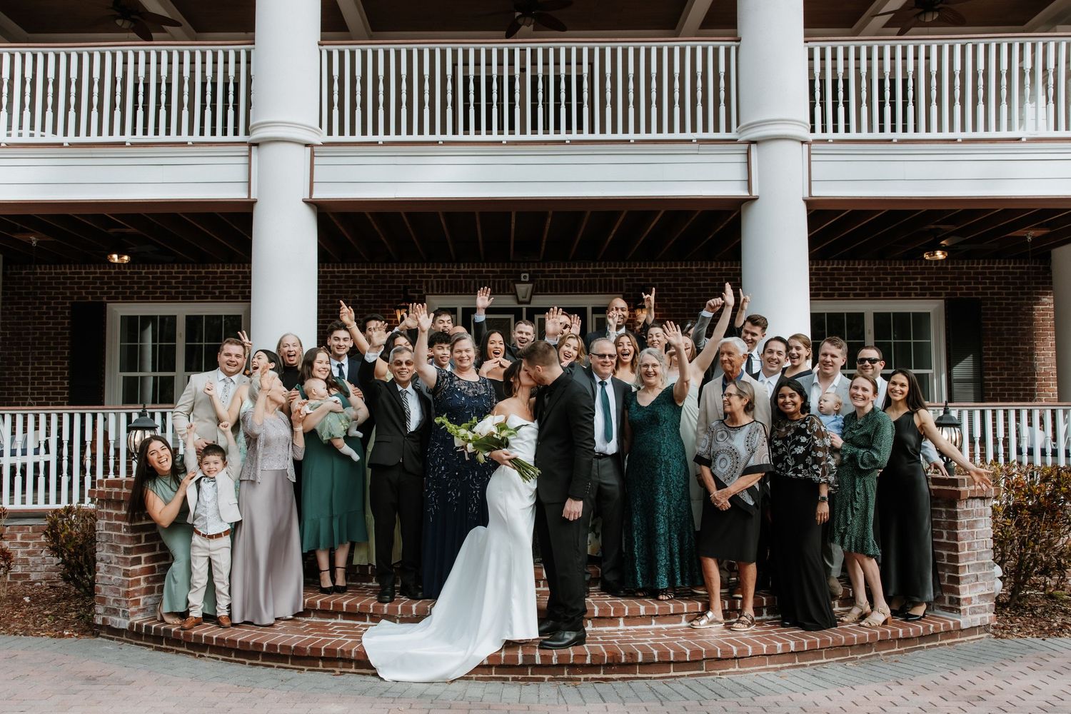 Large wedding party gathers for group photo on colonial-style porch with white columns and railing.