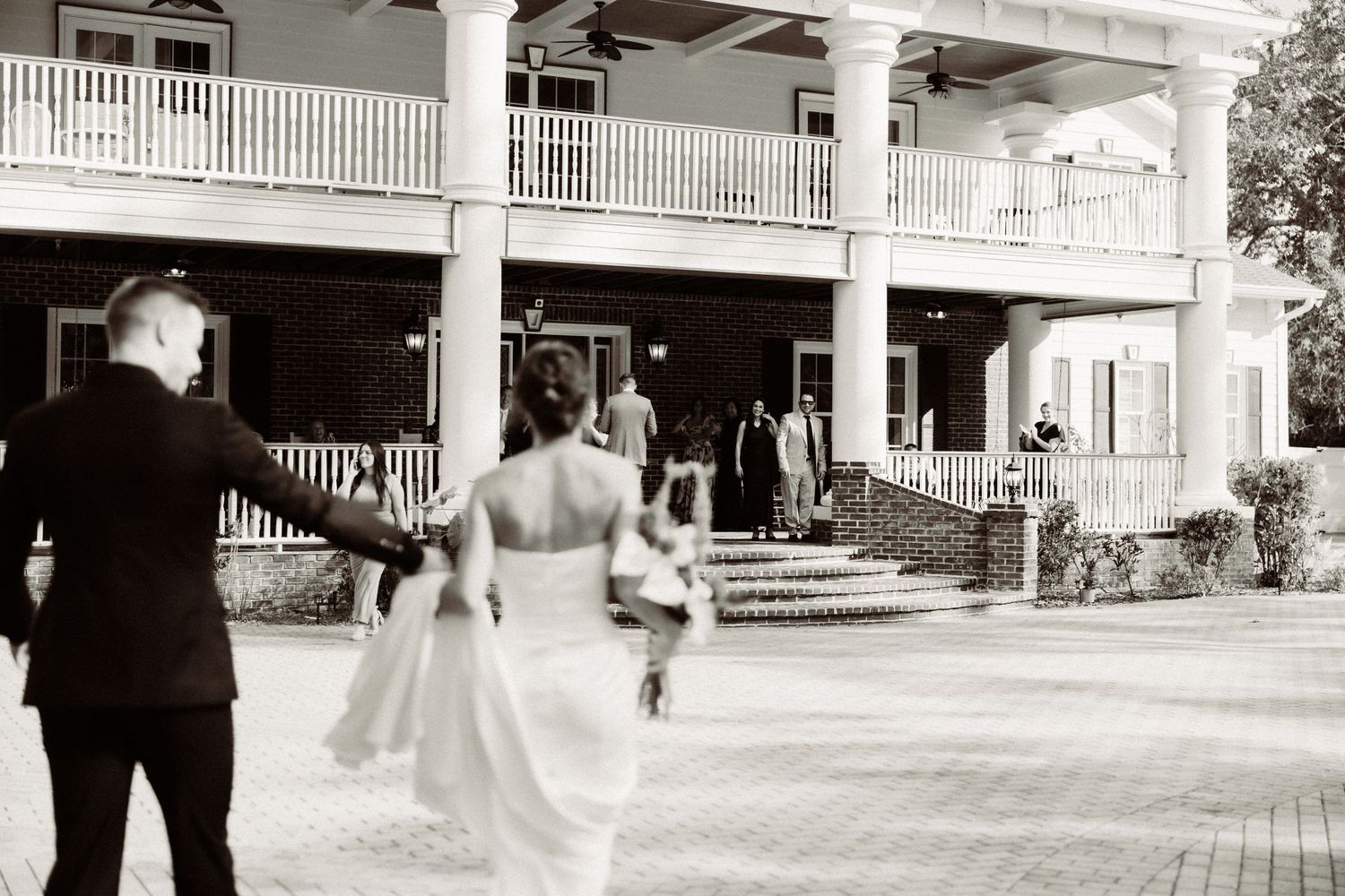 A couple approaches a grand white colonial style house with wrap-around porch during a daytime celebration.