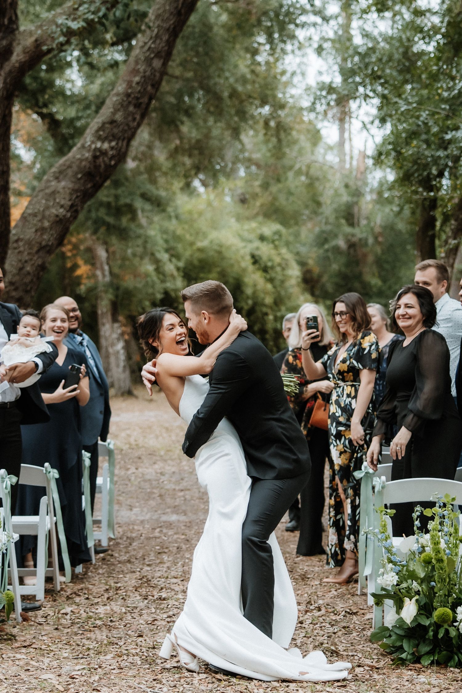 Wedding couple shares first dance on tree-lined path as guests capture the moment.