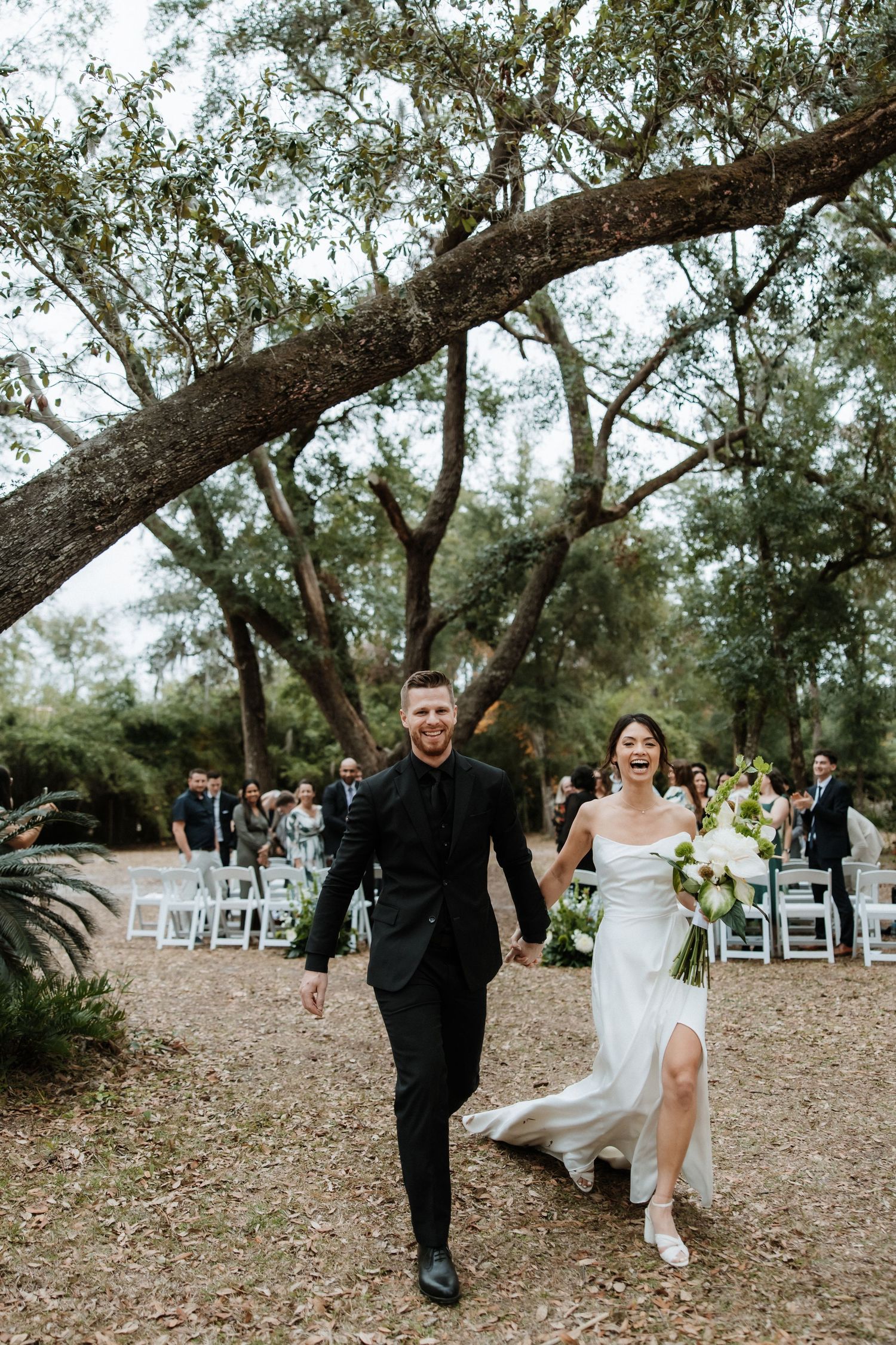 A couple walks down a rustic outdoor path after their wedding ceremony, with rows of white chairs visible in background.