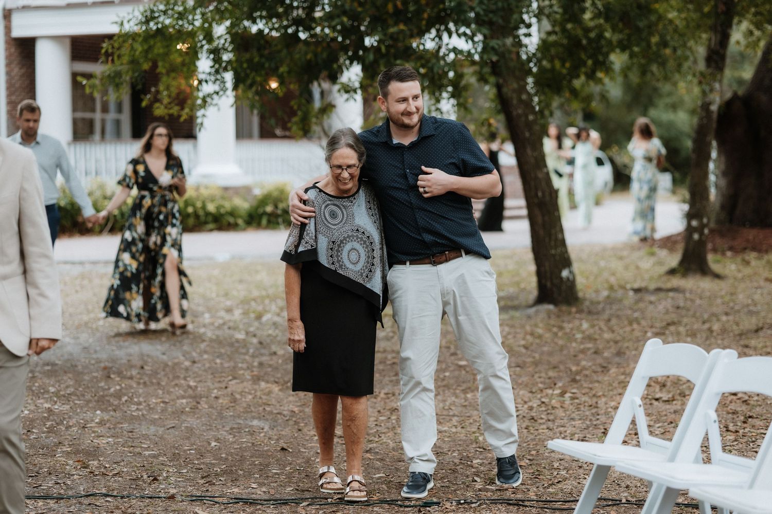 Guests gather outdoors at a wedding ceremony near white folding chairs under trees.