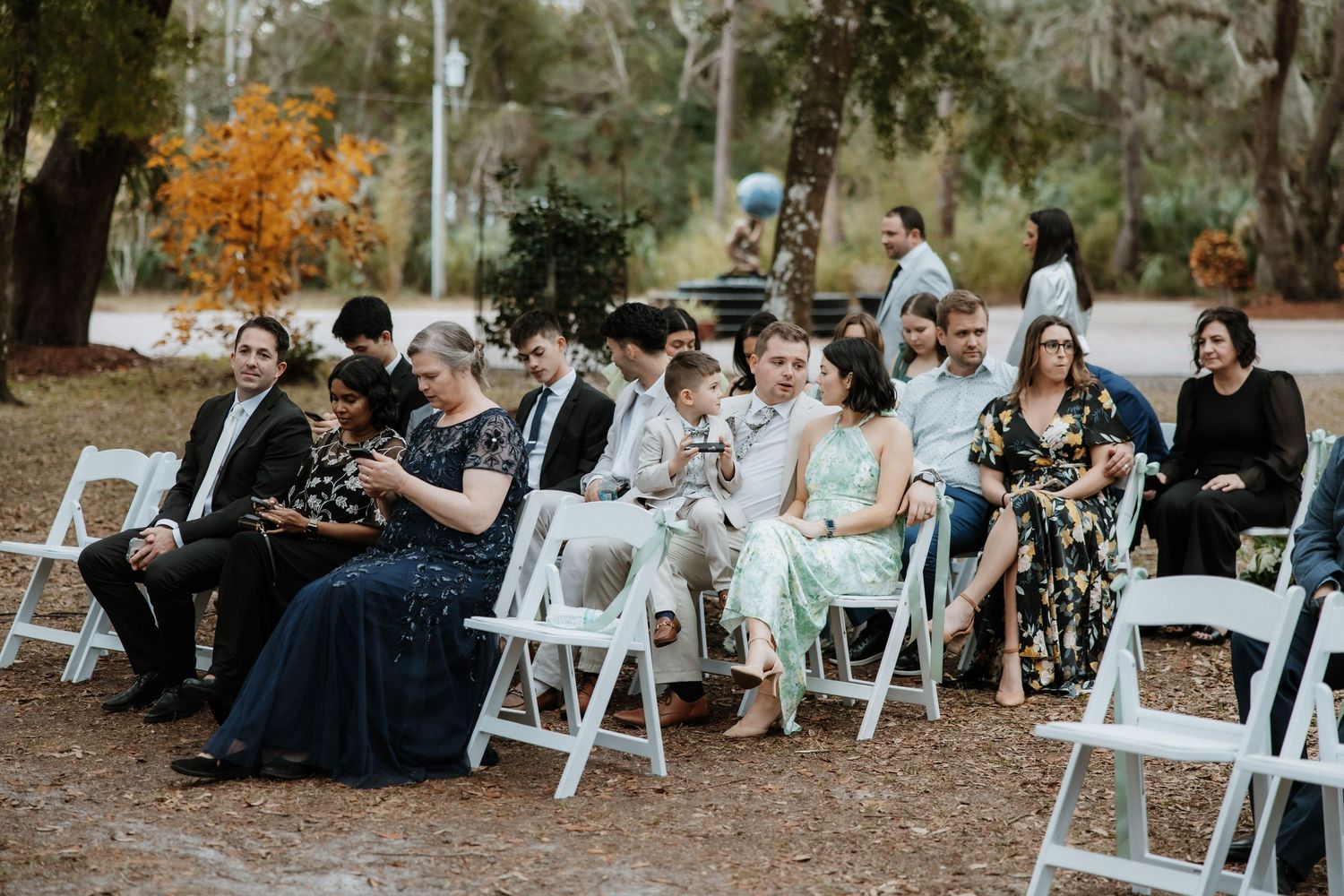 Wedding guests seated in white folding chairs at an outdoor ceremony in a wooded setting.