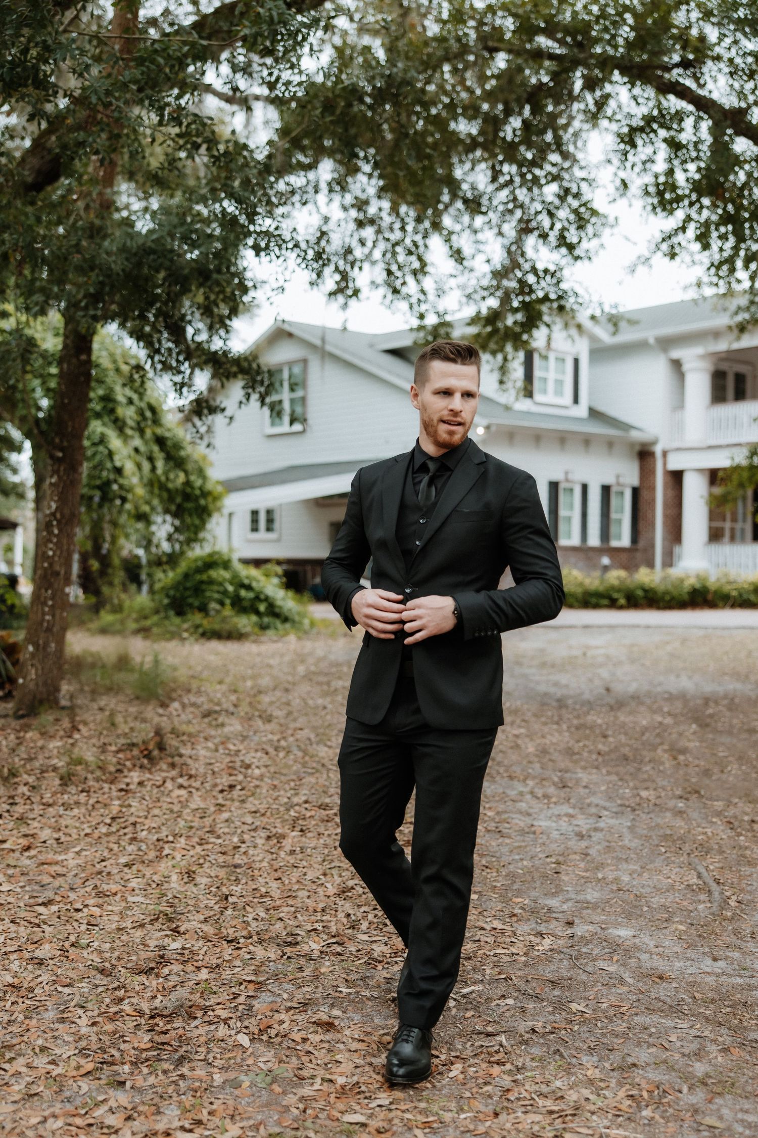 A person in an all-black suit walks on a leaf-covered path outside a white house with trees in the background.