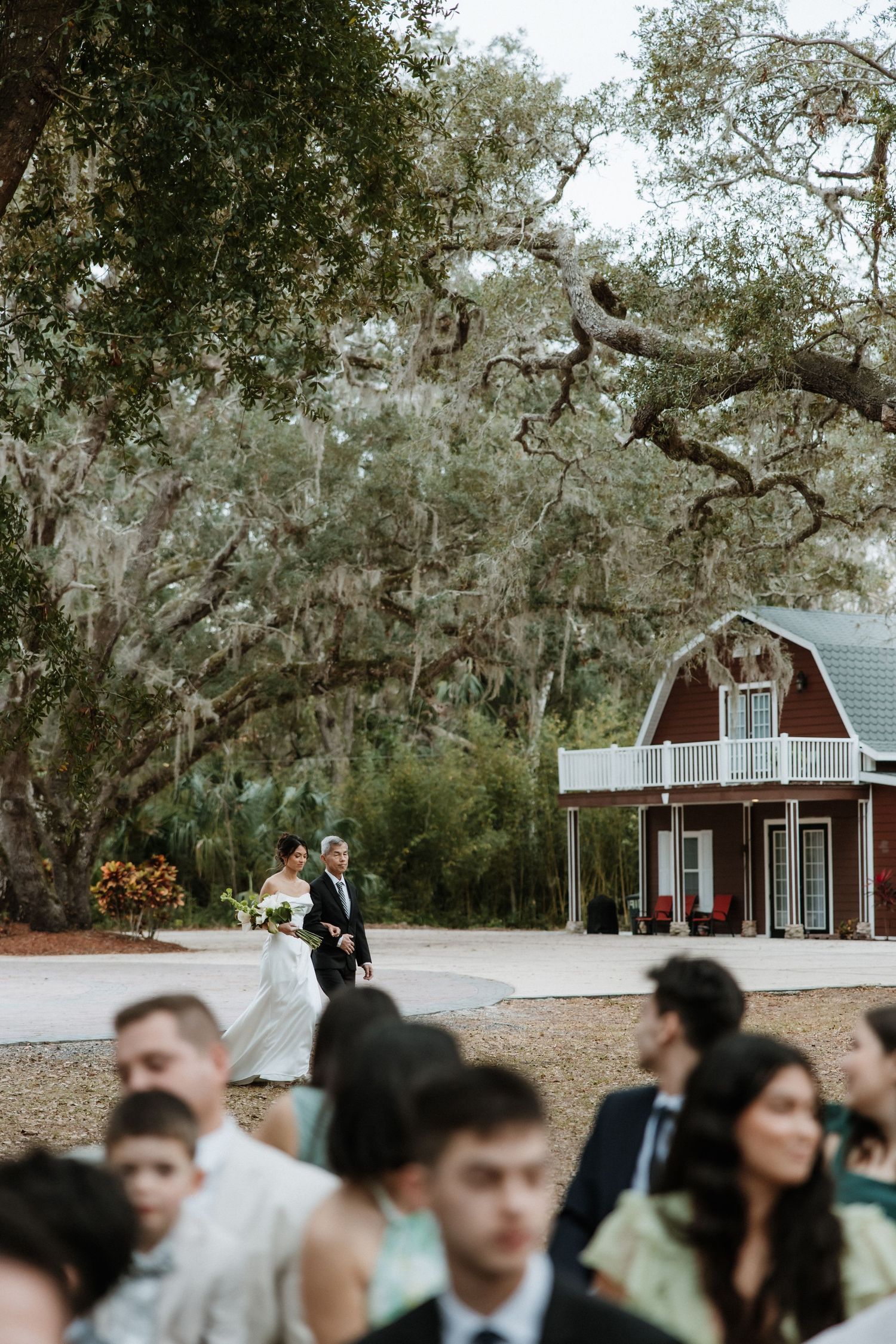 Outdoor wedding ceremony in front of a rustic red barn with oak trees draped in Spanish moss.