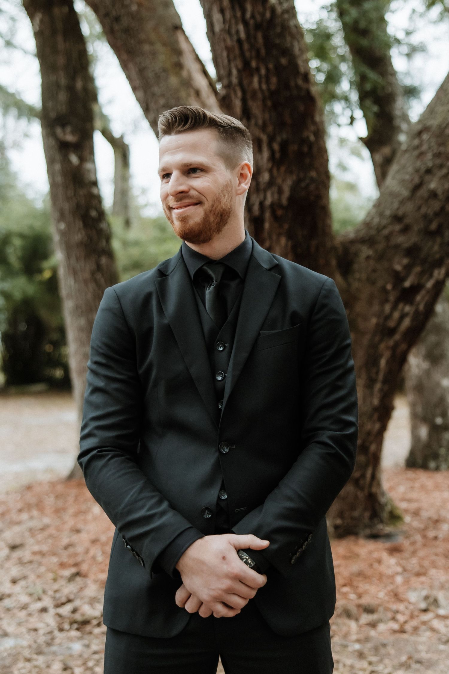 A groom in a black suit and tie stands confidently against a backdrop of trees.