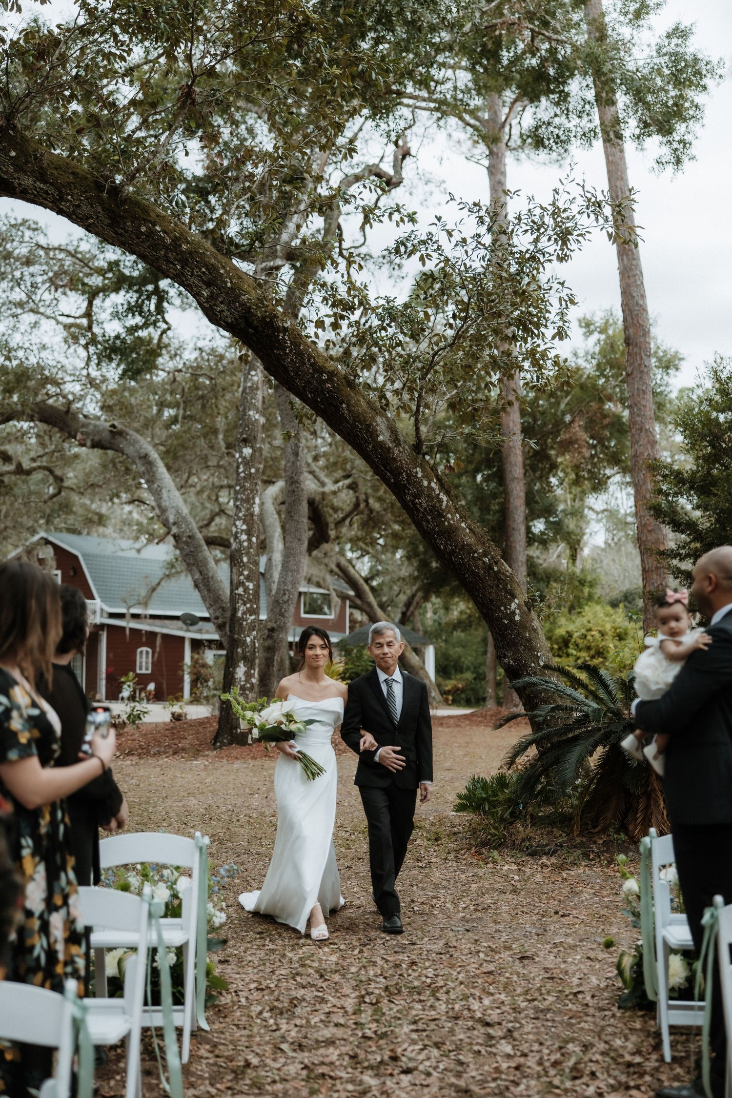 Outdoor wedding ceremony setup with white chairs nestled under large oak trees.