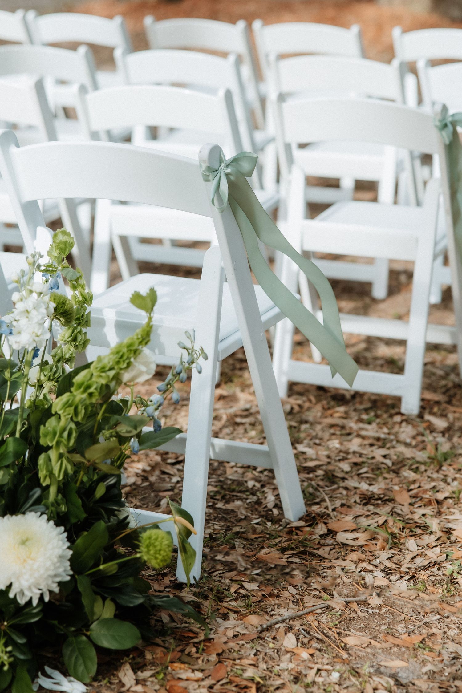 White folding chairs with sage green ribbons arranged outdoors with floral decorations for a wedding ceremony.