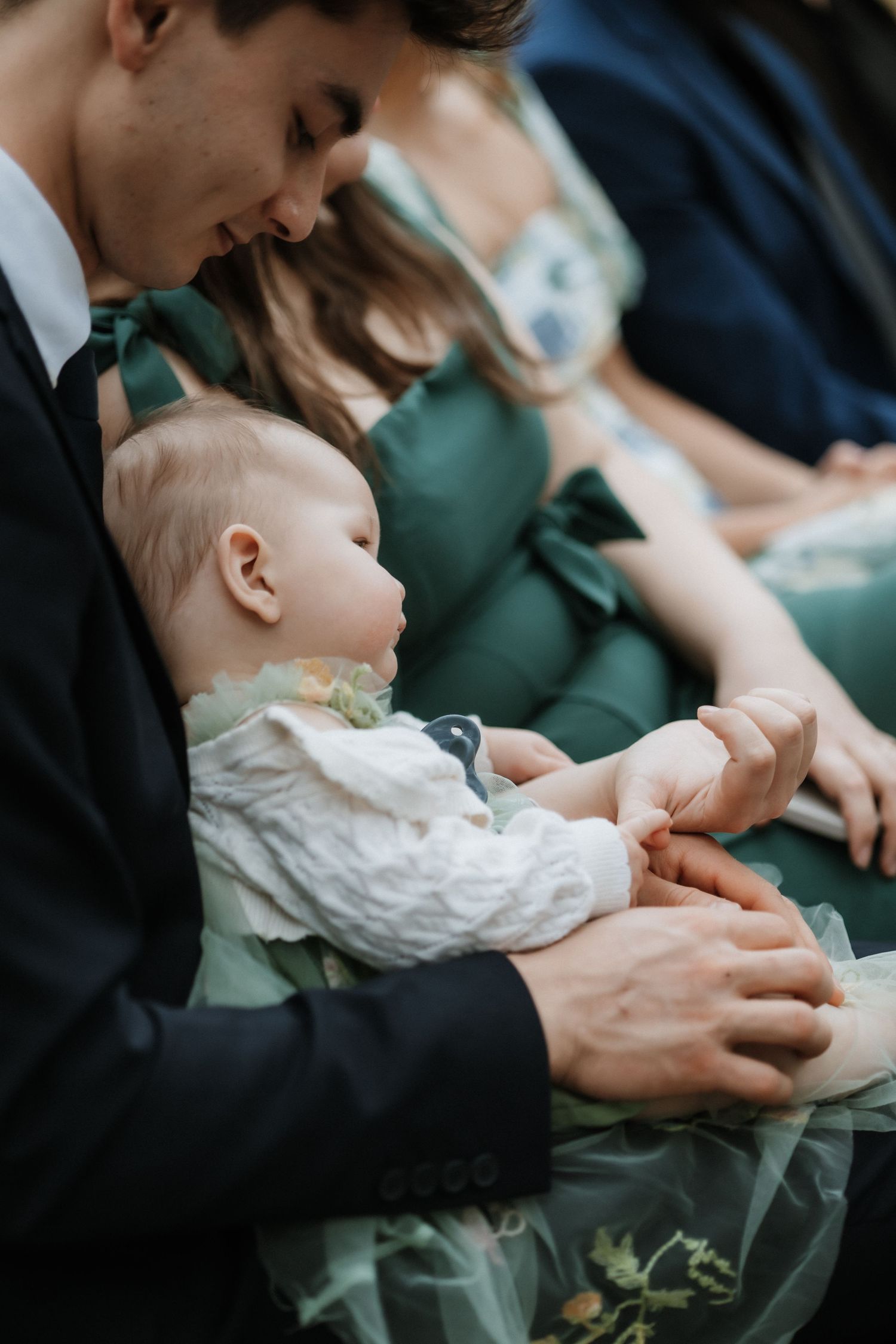 A tender moment shows a baby in white lace being held gently against a backdrop of emerald green formal attire.