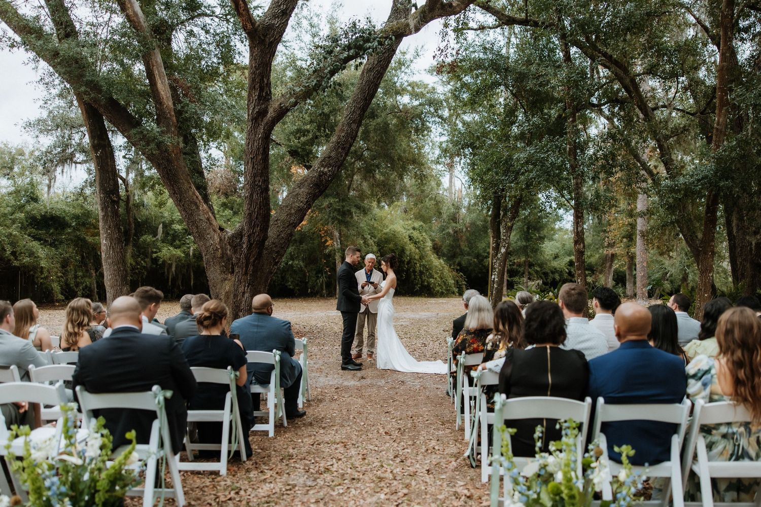 Outdoor wedding ceremony under oak trees with white chairs and floral aisle decorations.