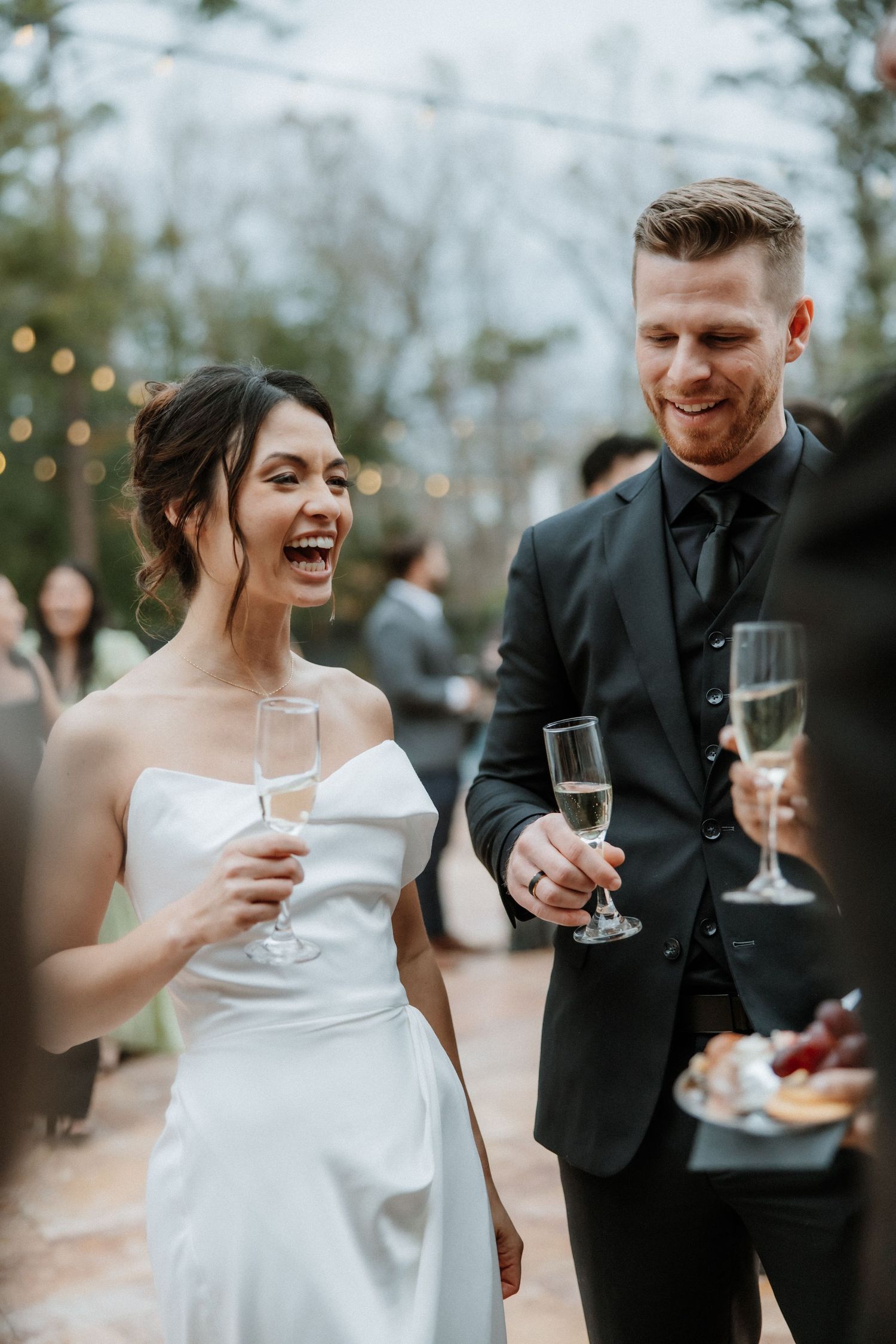 Joyful wedding couple sharing champagne and laughter during outdoor reception.