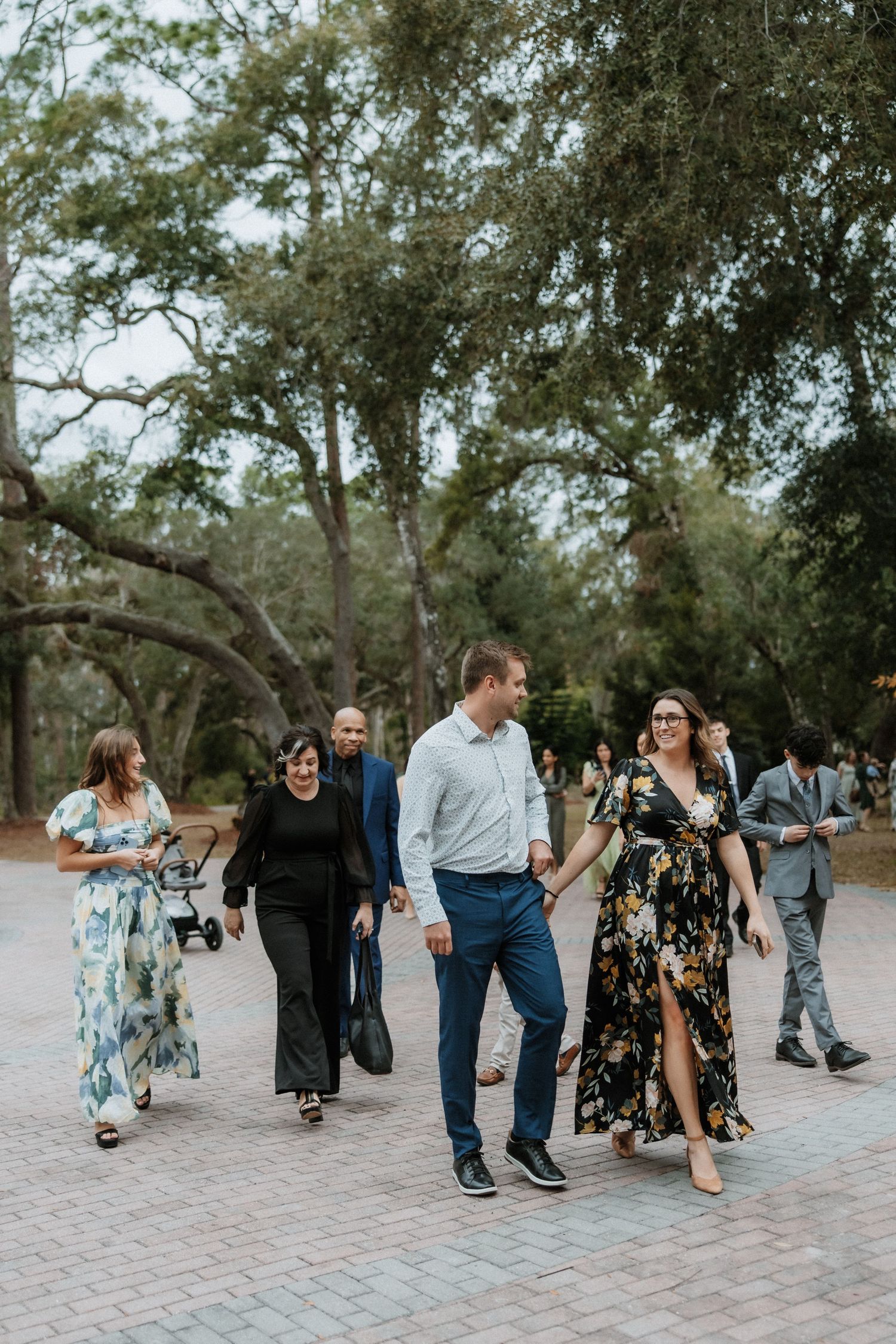 Wedding guests in formal attire walking along a brick pathway through a wooded area.