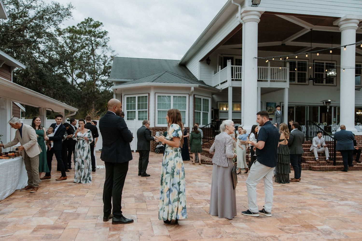Wedding guests gather on an outdoor patio area during a reception at a white colonial-style venue.