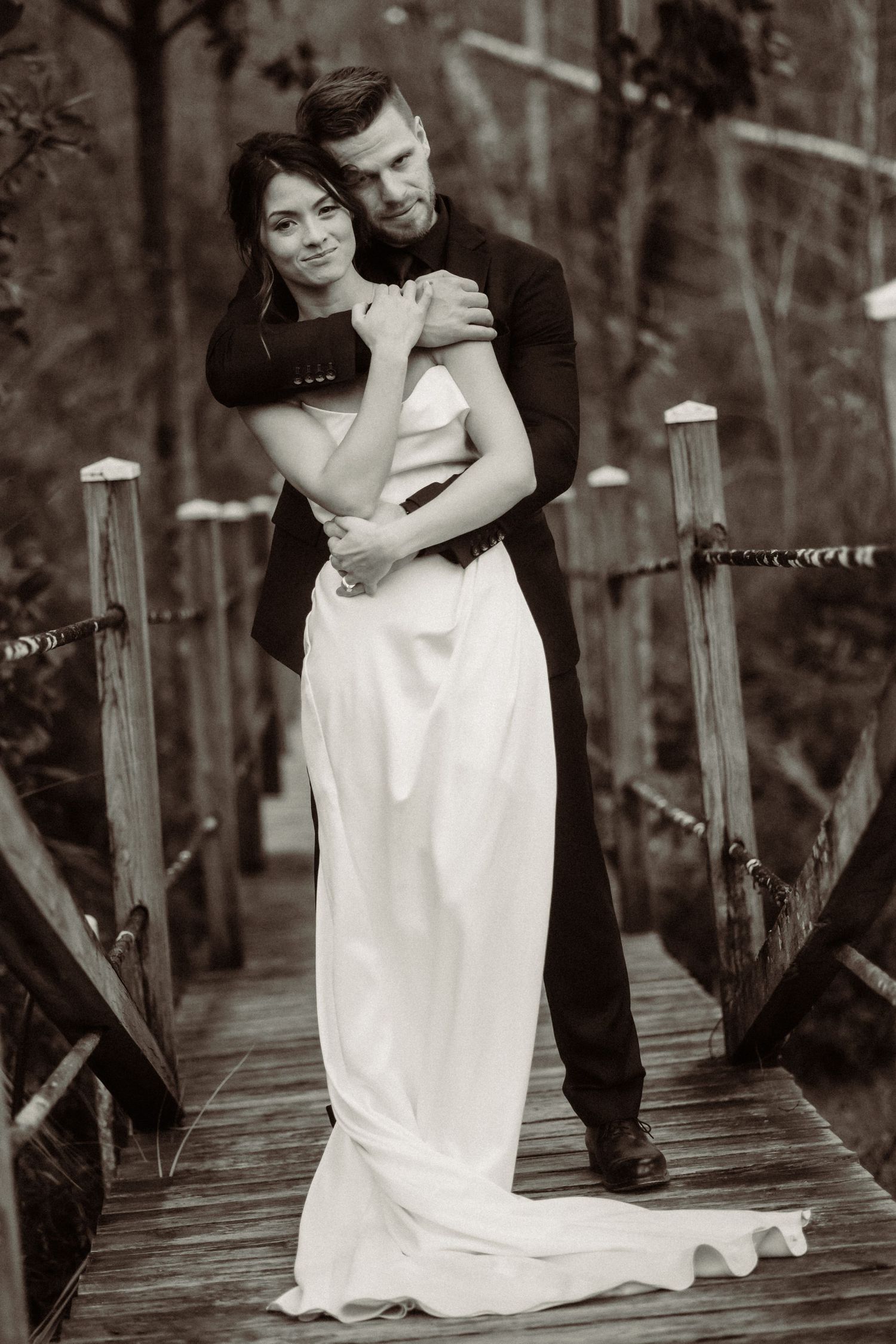 Classic black and white portrait of a couple embracing on a wooden bridge in formal attire.